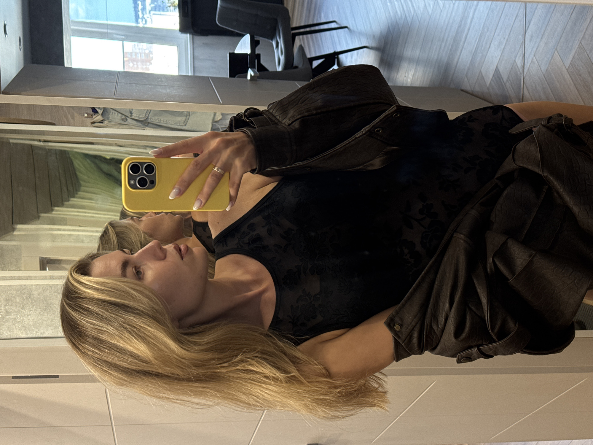 A blonde woman stands in front of a mirror, taking a selfie with her yellow smartphone. She is wearing a black, sleeveless, lace-patterned top and a dark brown leather jacket draped over her shoulders. Her hair is long and wavy, falling over her left shoulder. The mirror reflects the woman's profile, as well as parts of the room she is in. The room appears to be a modern interior with light-colored walls and a herringbone-patterned floor. In the background, visible in the mirror, is a window with a view of buildings and a cloudy sky, and a dining table with chairs.