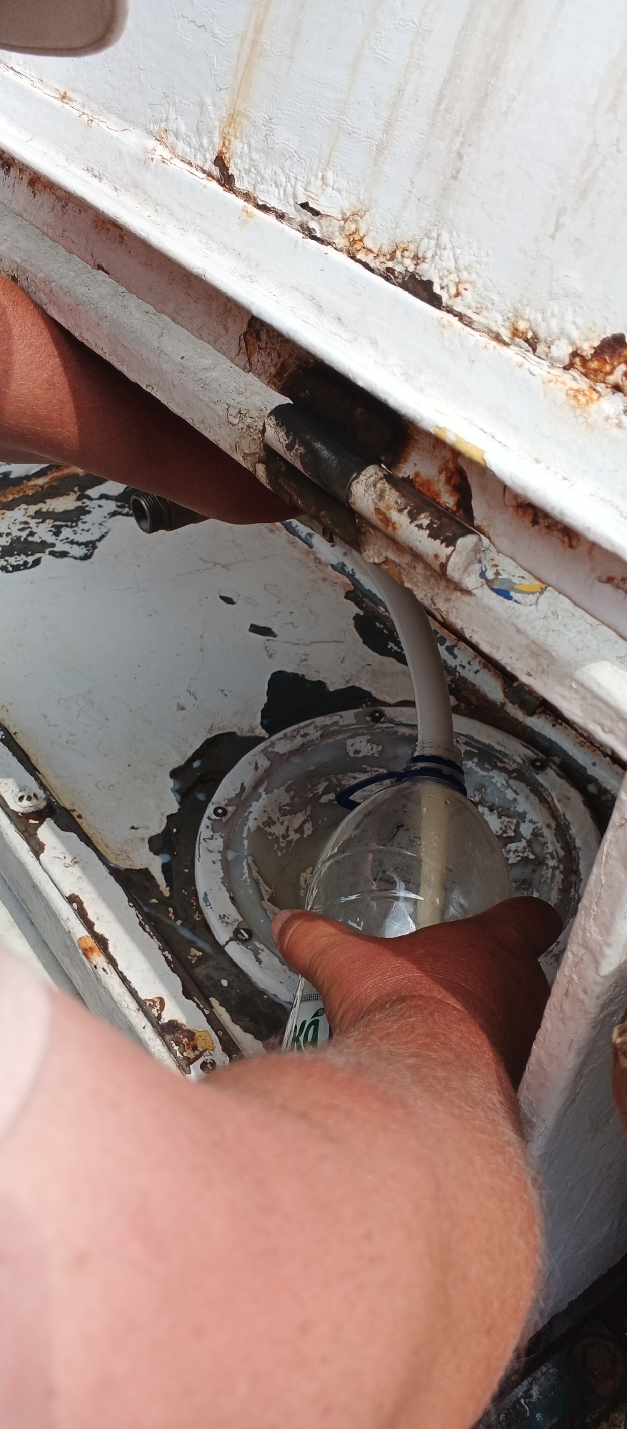 The image shows a close-up of a person's hands interacting with a white, rusted metal structure. One hand is holding a plastic bottle with a white tube inserted into its neck, which is being used to collect a light-colored liquid. The liquid appears to be dripping from the metal structure into a circular, metallic component below. The metal structure shows significant signs of rust and peeling paint. The scene is outdoors, with bright lighting suggesting it is daytime. The location in Smila, Ukraine, is not visually discernible from the image itself.