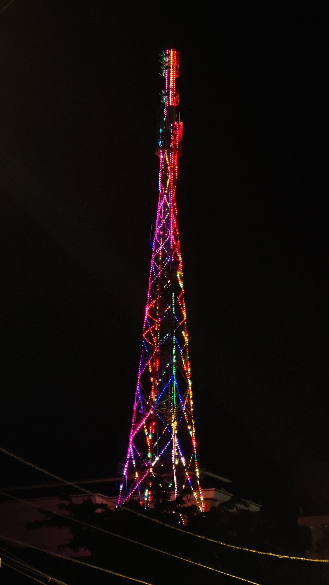 A brightly lit telecommunications tower stands illuminated against a black night sky. The tower is decorated with colorful lights in red, pink, blue, and green. Wires are seen in the lower left corner of the image. Below the tower, a few trees and parts of a building are barely visible in the dark. The scene is Yên Minh Commune, Vietnam.