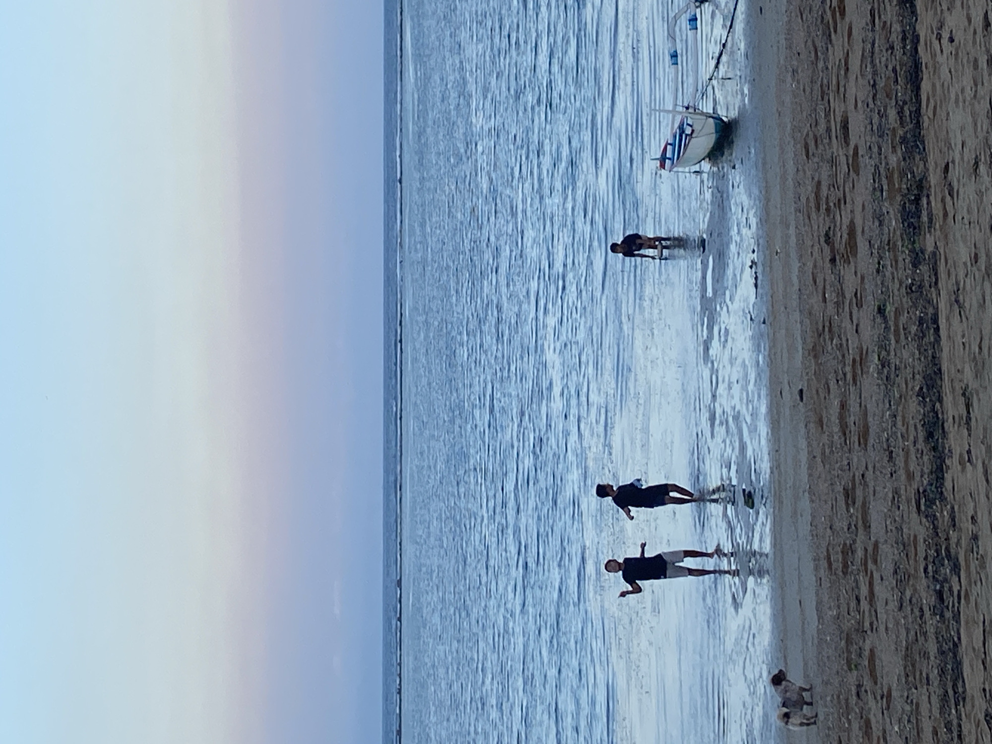 An aerial view of a beach in Denpasar, Indonesia, shows a calm ocean with a gradient of light blue to pastel pink in the sky. Three children, two in the foreground and one further back, are playing in the shallow water. A small dog stands on the sandy beach, looking towards the water. A white and blue boat is moored in the distance. The scene appears to be during sunset or sunrise, with soft lighting and a serene atmosphere.