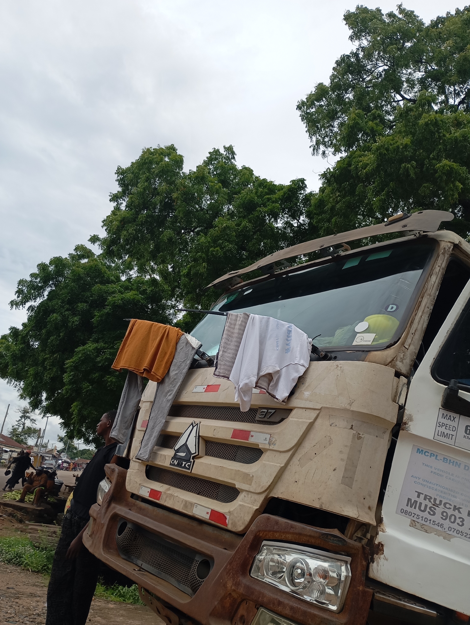 The image shows a large, weathered truck parked on a dirt road in Shendam, Nigeria. The truck's front is heavily damaged, with rust visible on the bumper and surrounding areas. Laundry, including an orange shirt and white t-shirt, is hung over the truck's grille and side mirror to dry. A young Black man in a black t-shirt and dark pants stands near the truck, looking to the left of the frame. In the background, other people are visible, some sitting on a wooden structure, and cars are parked along the road. Trees with dense green foliage line the background, and the sky is overcast. The overall impression is of a scene in a developing urban or semi-urban area where vehicles might be parked for extended periods and daily life activities, such as drying laundry, take place in proximity to transportation infrastructure.