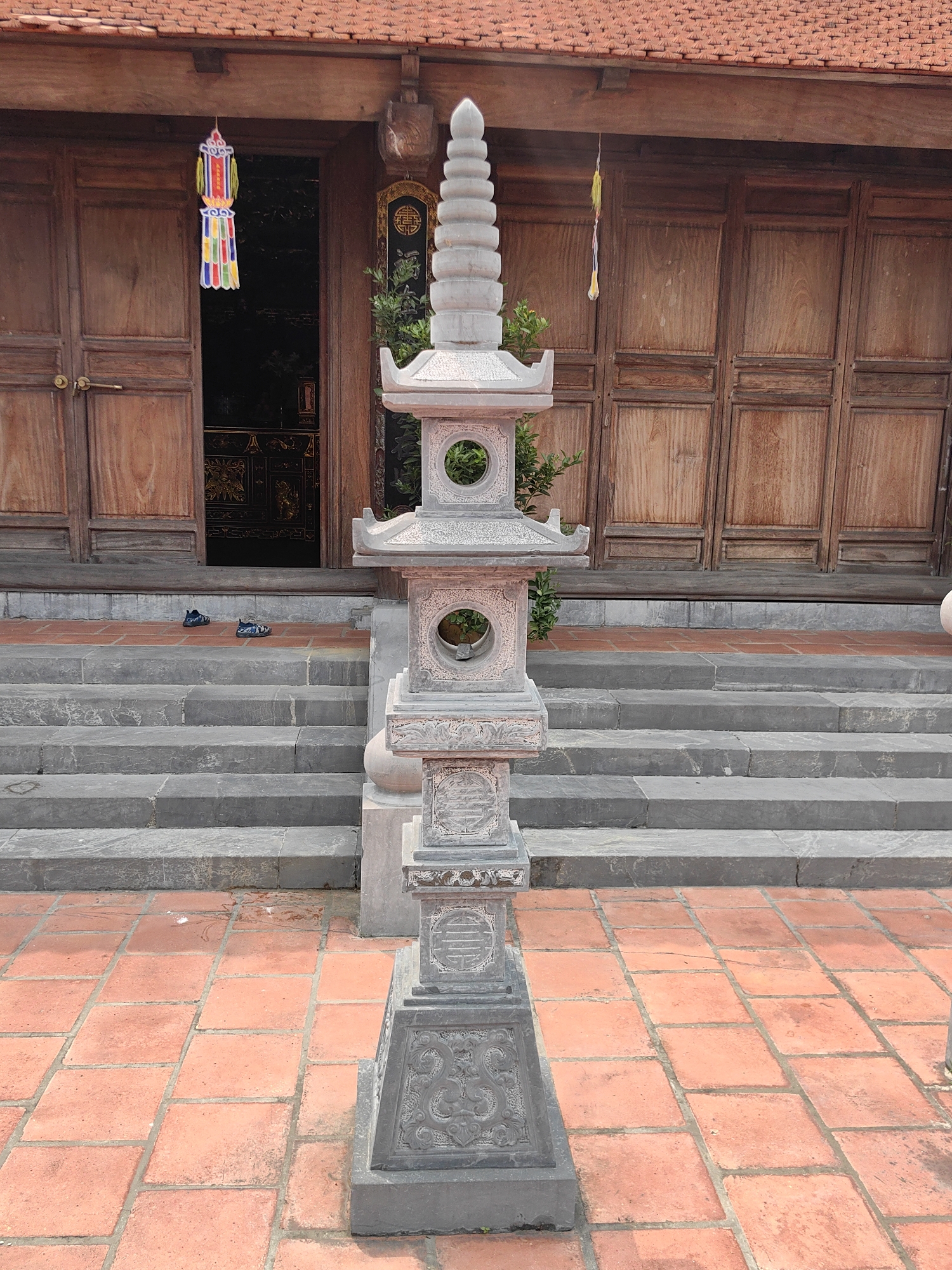 A stone pagoda stands prominently in front of a building in Hong Gai Ward, Vietnam. The pagoda, a multi-tiered structure with decorative carvings, is centrally located on a terracotta-tiled surface. Behind the pagoda, a set of gray stone steps lead up to a wooden building. The building has paneled wooden doors, some of which are open, revealing a glimpse of the interior. Hanging decorations are visible near the doorways. A pair of shoes sits at the base of the stairs, near the building's entrance. The architectural style suggests a temple or historical site.