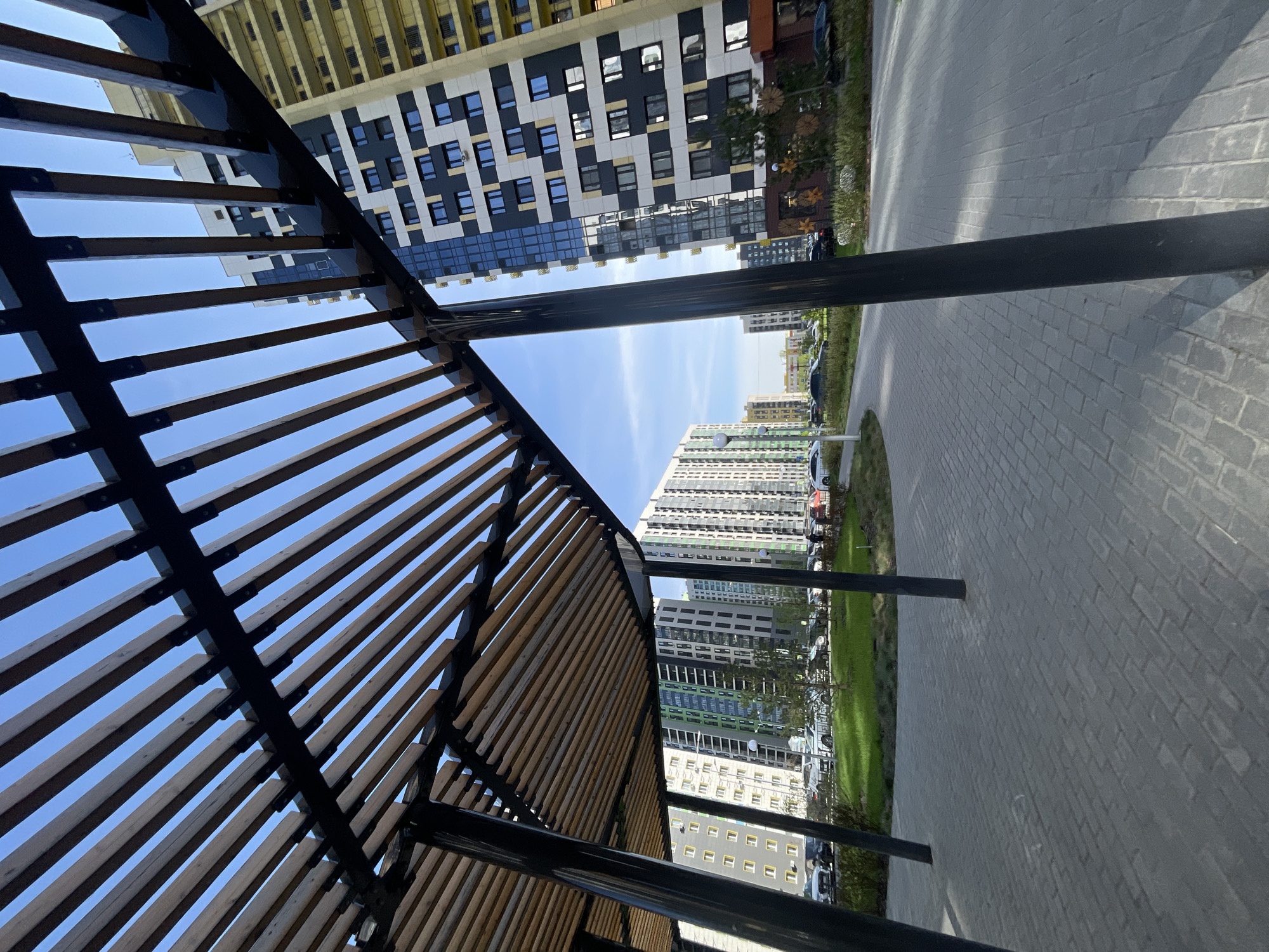 The image depicts an outdoor scene in Kazan, Russia, showing a modern urban landscape. A structure with a pergola-like design is in the foreground, featuring dark metal beams and light-colored wooden slats. The sky is visible through the slats. In the background are tall, modern apartment buildings with a mix of colors and architectural styles. A paved walkway runs alongside the buildings, and there's a strip of green grass and vegetation separating the walkway from the buildings. Some parked cars are visible. The image is taken from a low angle, looking upwards.