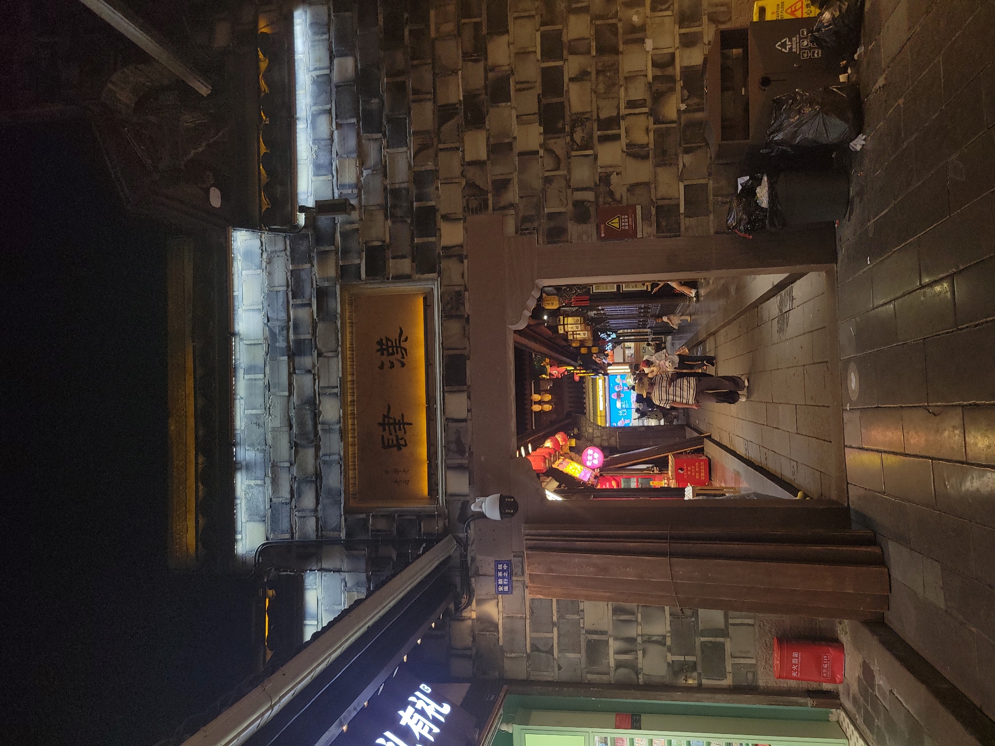 The image captures a nighttime scene in Wuhou District, China, likely a pedestrian walkway lined with traditional architecture. A prominent brick wall, possibly part of an ancient structure or a themed development, features a horizontal sign with Chinese characters. Below this sign, an arched entryway leads into a dimly lit area adorned with red lanterns and further architectural details. A CCTV camera is mounted on the wall near the entrance. On the left side, a storefront with illuminated signage is visible, and a red fire extinguisher is placed on the ground. The walkway is paved with stone slabs and shows reflections from overhead lighting. Several individuals are walking through the archway and along the path, their activities not clearly discernible due to the low light. Trash receptacles and overflowing plastic bags are situated on the right side of the walkway, adjacent to the brick wall. The overall atmosphere suggests a historic or cultural district open for evening strolls and commerce.