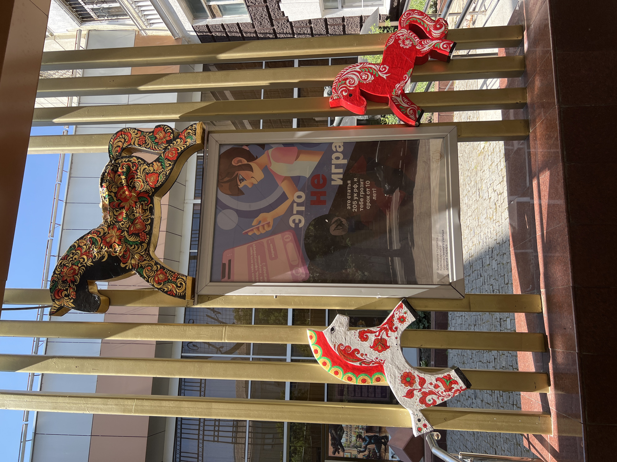The image shows an outdoor scene in front of a building, likely in Krasnodar, Russia, due to the presence of a poster with Russian text and an inscription referencing a student from Krasnodar. Three decorative wooden horses are mounted on golden vertical poles. The largest horse, at the top, is black with ornate red, gold, and green floral patterns. To the left, a smaller white horse with red and green floral designs is mounted on a pole. To the right, a smaller red horse with white and green floral patterns is also attached to a pole. Between the horses, a framed poster is displayed. The poster features an illustration of a young person holding a smartphone, with Russian text on it. The background shows a building with windows and a blue sky. The lower part of the image reveals a patterned tiled floor and a red tiled base.