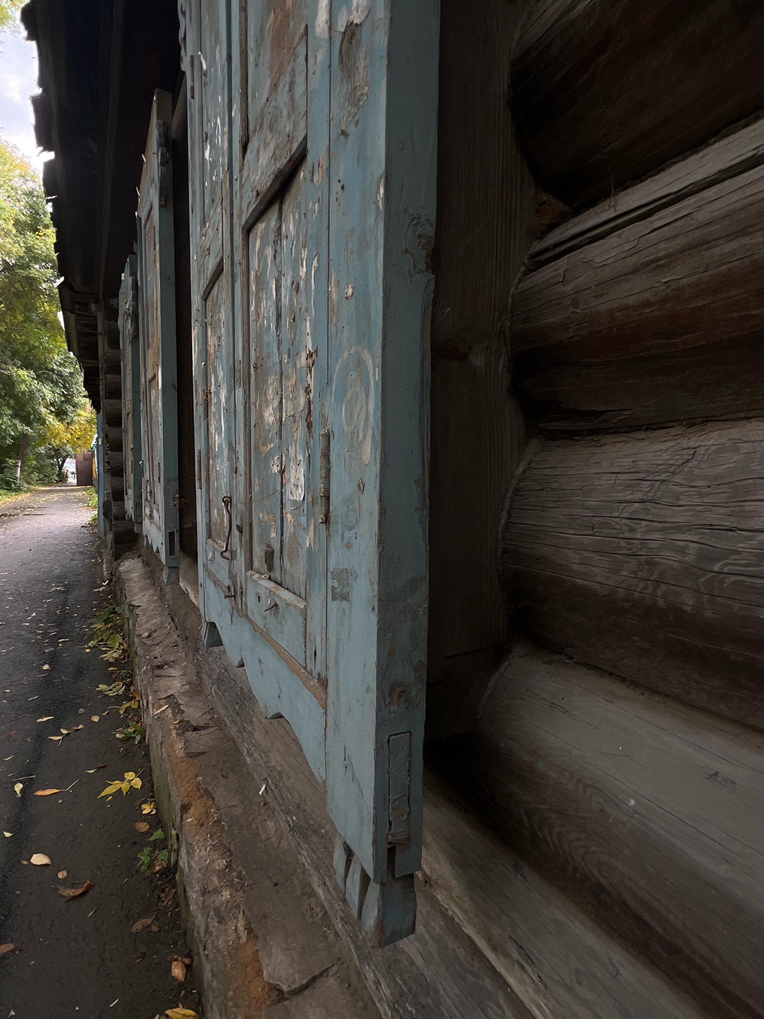A low-angle, close-up, outdoor, medium shot shows the weathered, light blue painted wooden shutters of an old wooden house, with peeling paint and visible rust on the hinges. The shutters are partially open, revealing a dark interior. To the left of the shutters, an asphalt path is visible, with fallen yellow and brown leaves scattered on it. Beyond the path, a line of trees with autumn foliage forms the background. To the right of the shutters, the rough, horizontal logs of the house's exterior are visible, with deep grain and a dark, weathered appearance. The base of the house features a stone foundation that is partially covered by dirt and leaves. The scene appears to be in a rural or historical part of Yekaterinburg, Russia, given the architectural style. No people are present in the image.