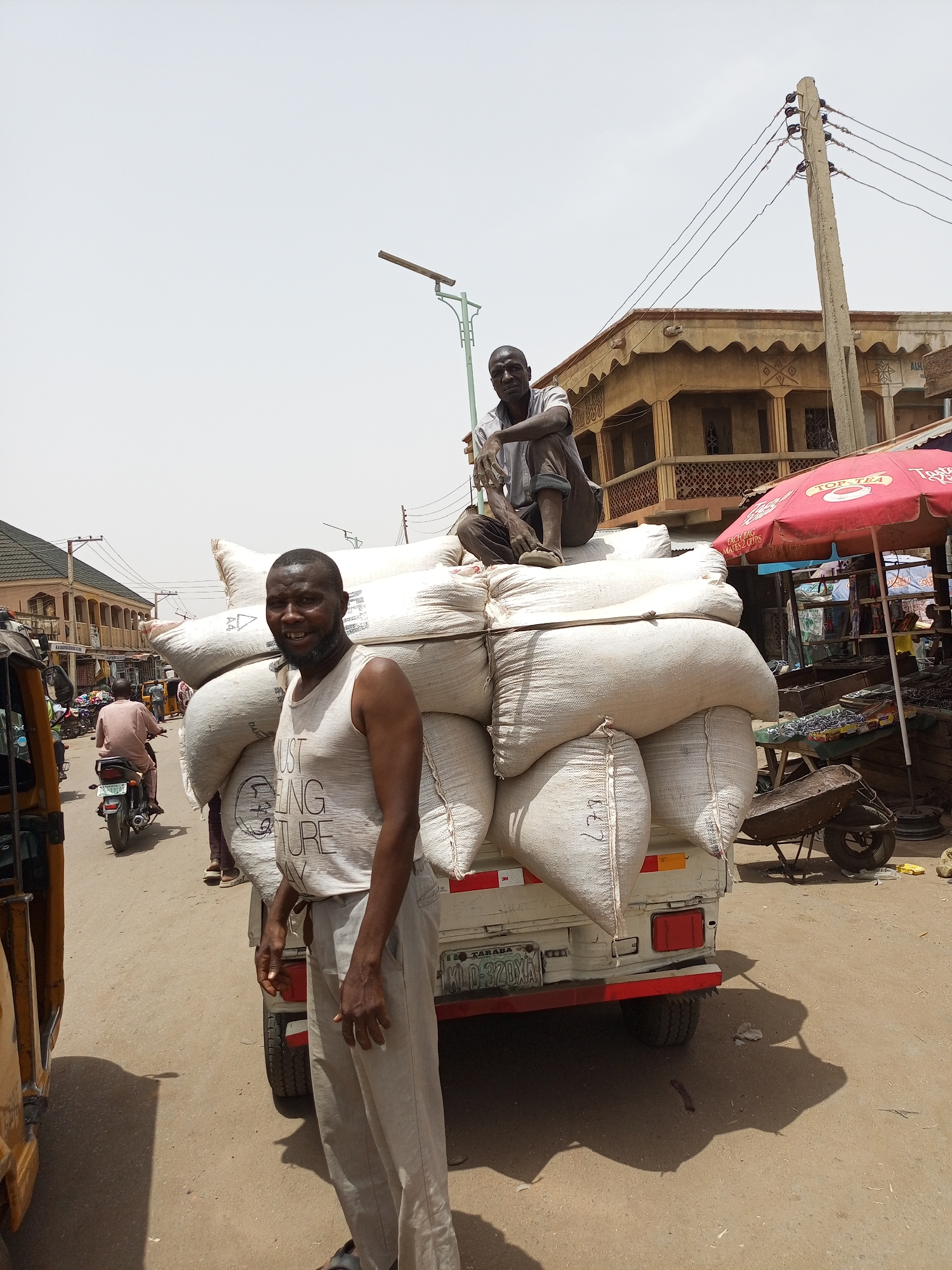 The image depicts a street scene in Arawa, Nigeria. In the foreground, a man stands beside a small truck heavily laden with white sacks. A second man sits atop the sacks. The man in the foreground is wearing a white tank top with dark text and light grey trousers. The man atop the sacks is wearing a light-colored shirt and dark trousers. The truck's license plate reads "TARABA D-320XA". The street is dusty and shows signs of traffic. Behind the truck, there are other vehicles including a motorcycle and a yellow tricycle taxi, and pedestrians are visible. A building with a decorated facade is in the background to the right. A red umbrella is also visible in the background, providing shade for a market stall. A utility pole stands to the right of the building.