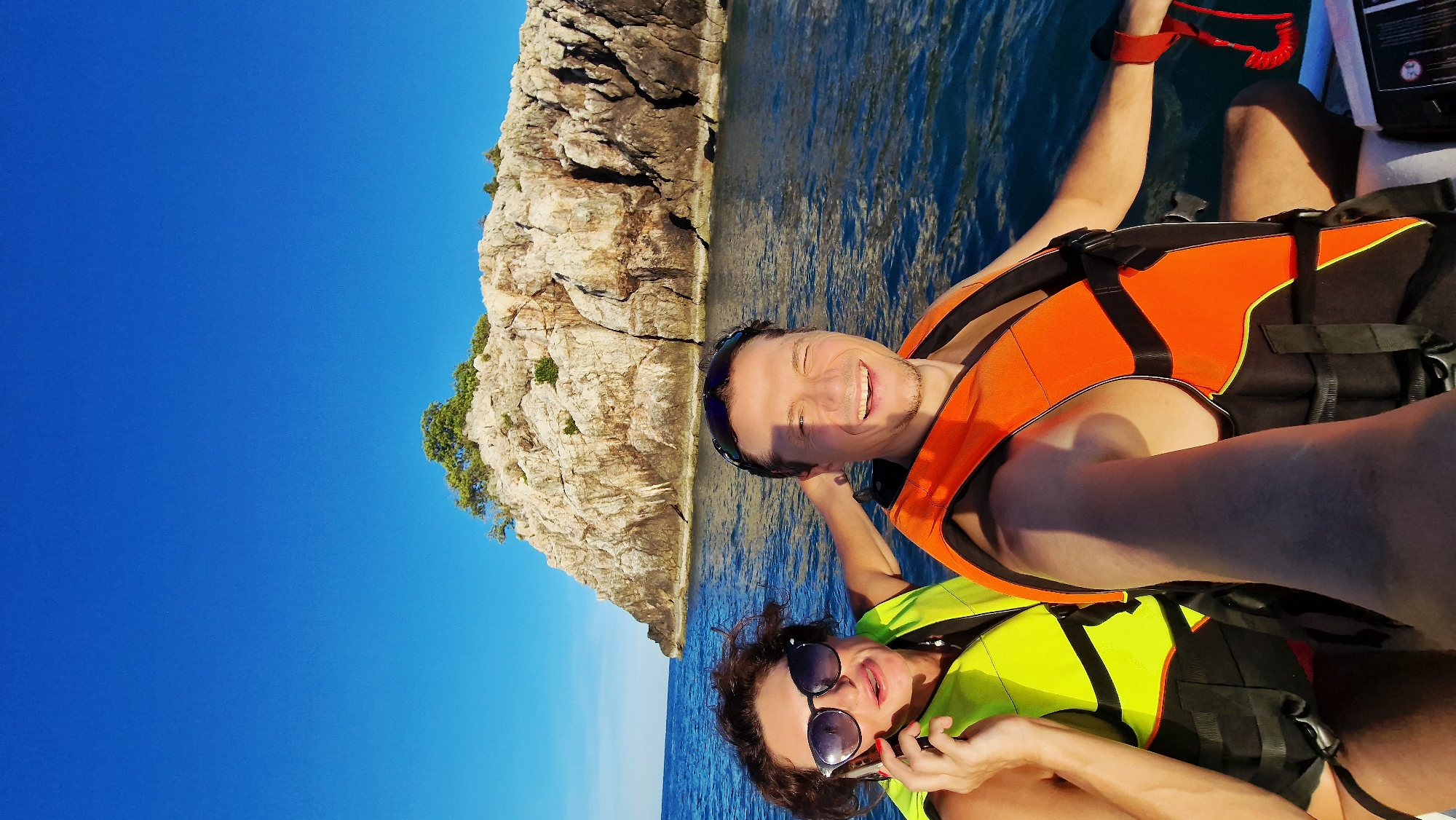 This is a selfie-style photo of a man and a woman on a boat in the ocean, with a rocky island in the background. The man in the foreground is wearing an orange life jacket and sunglasses pushed up on his head, smiling widely and looking at the camera. The woman, to his left, is wearing a yellow-green life jacket, sunglasses, and holding a smartphone to her ear. She is looking off to the side with a surprised expression. The water is blue and choppy, and the sky is clear and bright blue. The island in the background is rocky and has some sparse vegetation on top. The scene is likely a tourist activity in Choeng Mon, Thailand, known for its beautiful beaches and opportunities for water sports and island hopping.