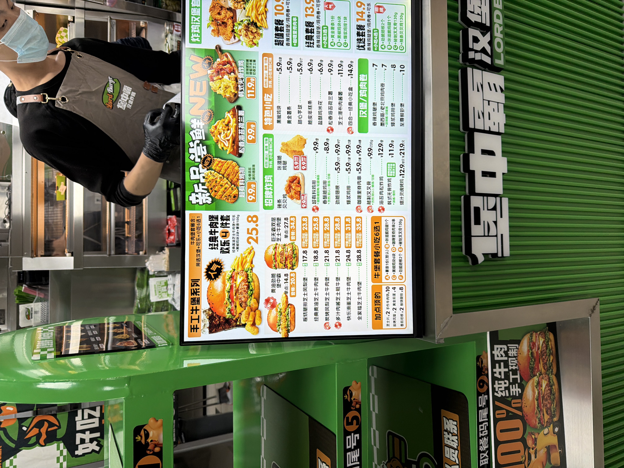 A masked person in a black long-sleeved shirt and brown apron stands in front of a Lord Burger restaurant menu in Jiangning District, China. The menu features a variety of burgers, chicken, and snacks. The menu is on a green wall. The menu boards are displaying both Chinese and English.