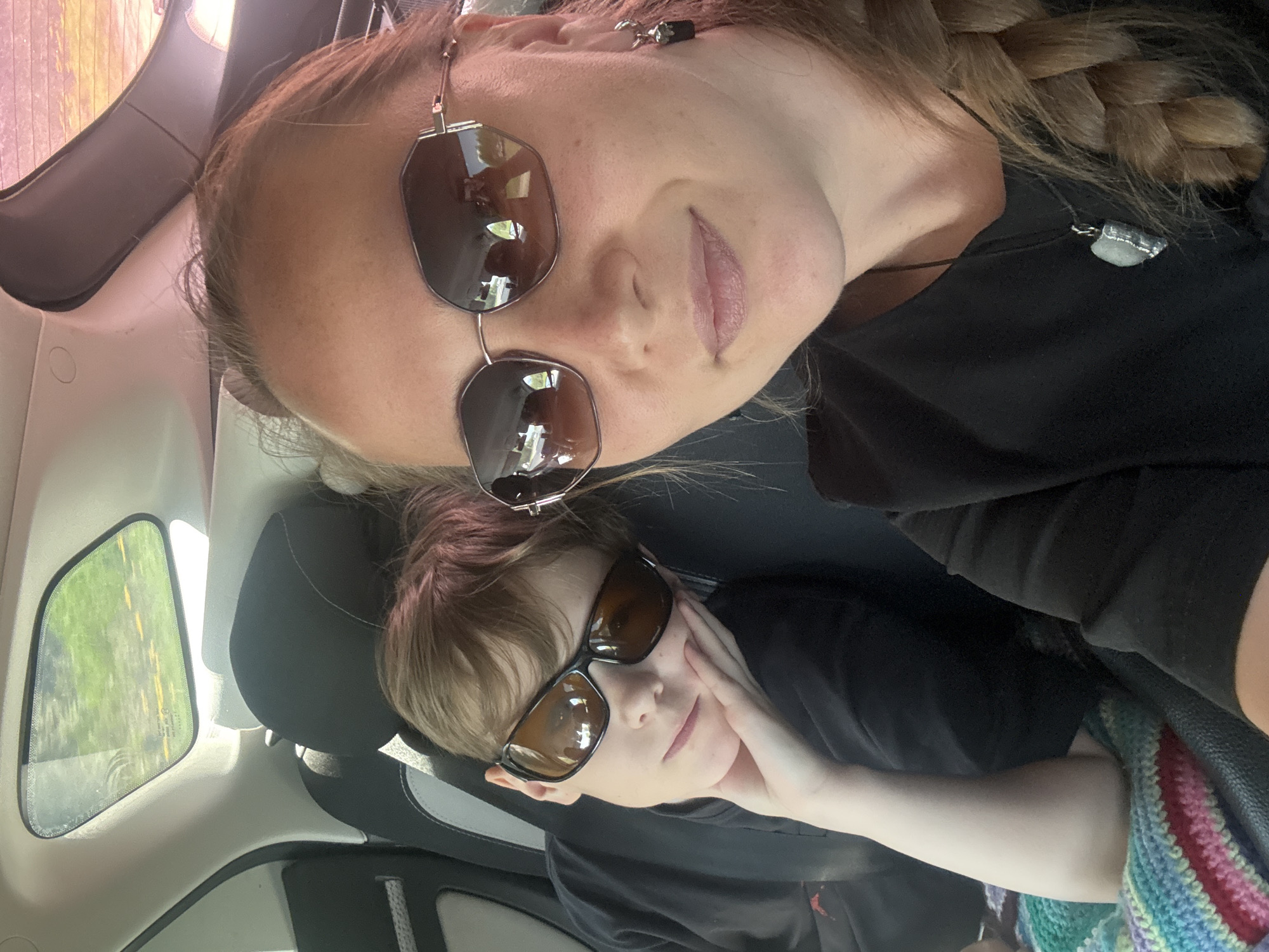 Two people are in a car. The person at the top of the image is wearing sunglasses and has a braid. The person at the bottom of the image is also wearing sunglasses and resting their head on their hand. The background shows the interior of a car, including seats and a window. The location is not discernible from the image.