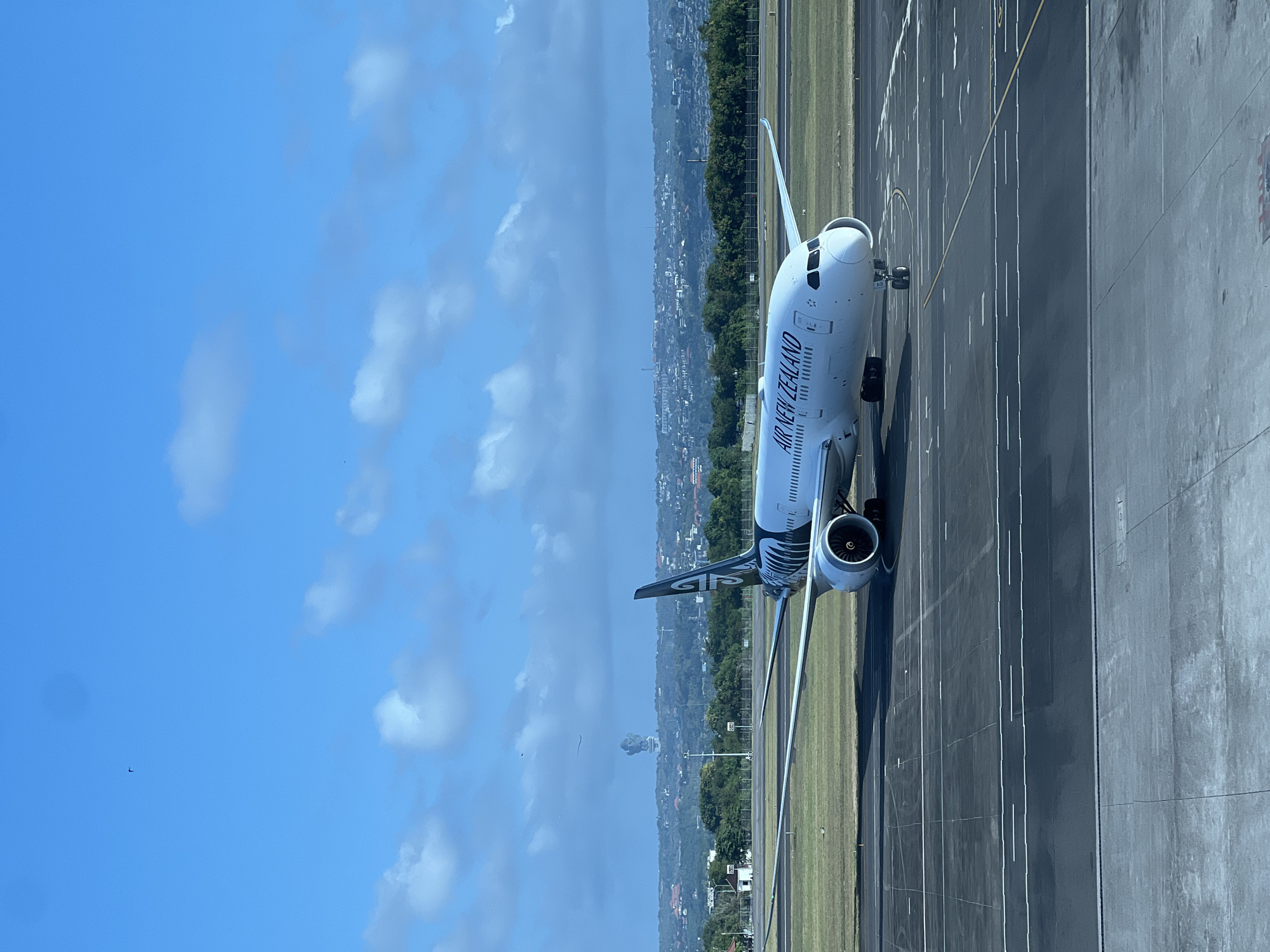 An Air New Zealand Boeing 787-9 Dreamliner aircraft is parked on the tarmac at Ngurah Rai International Airport (DPS) in Denpasar, Bali, Indonesia, near Kuta. The aircraft, bearing the registration ZK-NZB, is predominantly white with black markings and the distinctive Air New Zealand logo on its tail. It is positioned on a paved runway, with various painted lines indicating taxiways and lanes. In the background, a hazy cityscape with buildings and lush green trees stretches across the horizon. Above the city, a large statue of Garuda Wisnu Kencana is faintly visible through the atmospheric haze. The sky is a clear blue, dotted with scattered white clouds. The foreground shows a section of the airport terminal building's glass facade, suggesting the photo was taken from inside the airport. There are no people visible in the image.