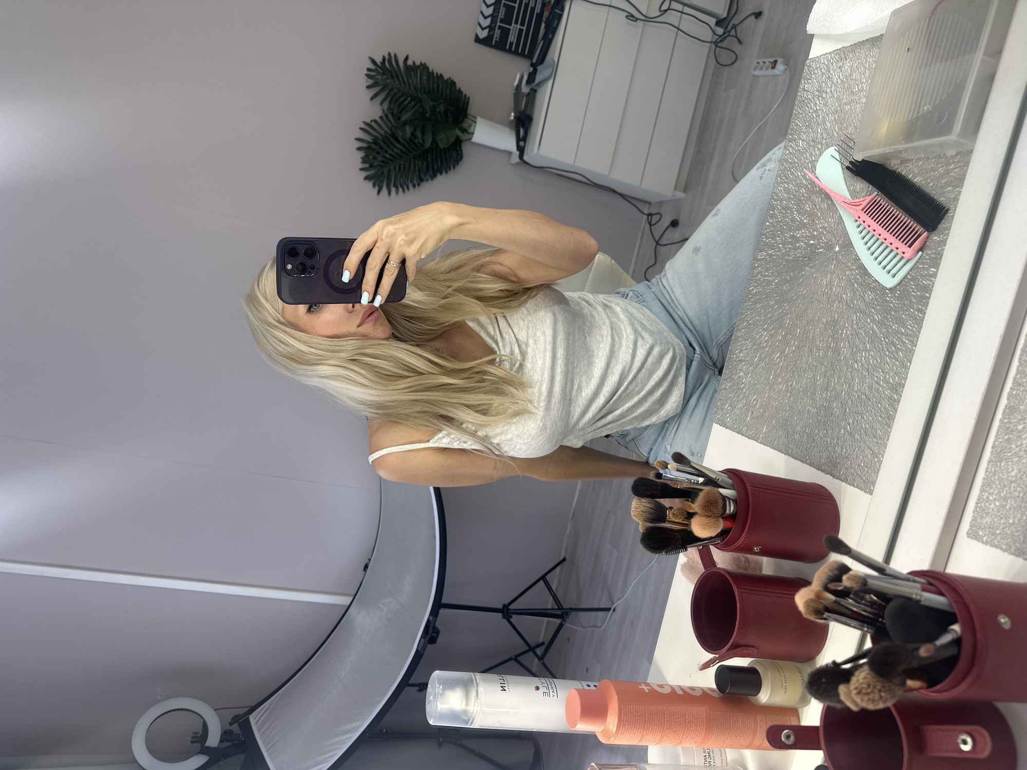 A woman with blonde hair is taking a selfie in a mirror. She is wearing a grey tank top and light blue jeans. The woman has light blue nail polish on her fingernails and is wearing a ring on her right hand. She is holding a black smartphone up to take the photo. The background appears to be a room with a vanity or makeup station. There are several makeup brushes in red containers on the vanity, along with some bottles of beauty products. A ring light and a softbox are visible in the reflection of the mirror, suggesting a well-lit setup for filming or photography. Two combs, one pink and one black, are also visible on the vanity. In the background, there is a white cabinet and a decorative plant. The overall scene suggests a personal grooming or content creation space. No time of day or weather information can be determined. No emotions are clearly discernible from the image. Text is visible on bottles of beauty products, but the text is mirrored and not fully legible.