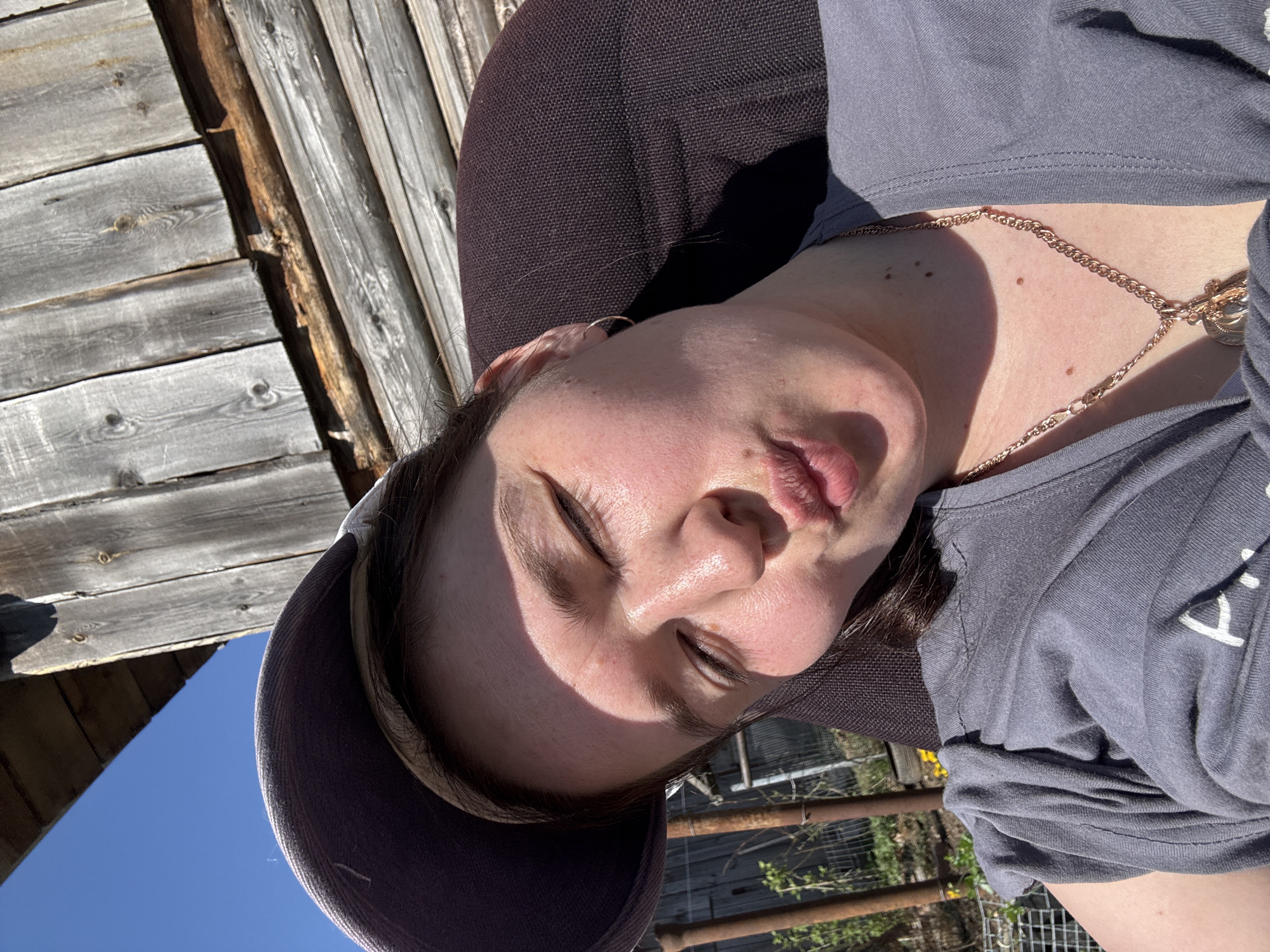 A woman is lying back, possibly in a chair, outdoors on a sunny day. She is wearing a baseball cap and a gray t-shirt with a gold chain. In the background is a wooden structure that appears to be a building or fence. Beyond that, there is a glimpse of blue sky. There is a metal fence and some greenery in the blurred background. The image does not contain enough information to ascertain the location in the city of Фоки, Russia.