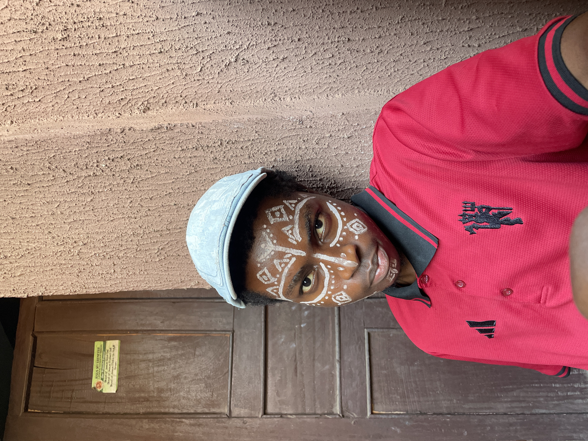 A young, dark-skinned person with white tribal-style paint on their face poses for a portrait, wearing a light blue hat and a red polo shirt with black accents. The shirt has an embroidered design near the collar. They are standing in front of a brown wooden door, with a label on the door that includes the text "JESUS MY EBENEZER" and mentions "Blood of Jesus." The environment appears to be a simple, textured wall. The scene seems to be outdoors in Nkpor, Nigeria.