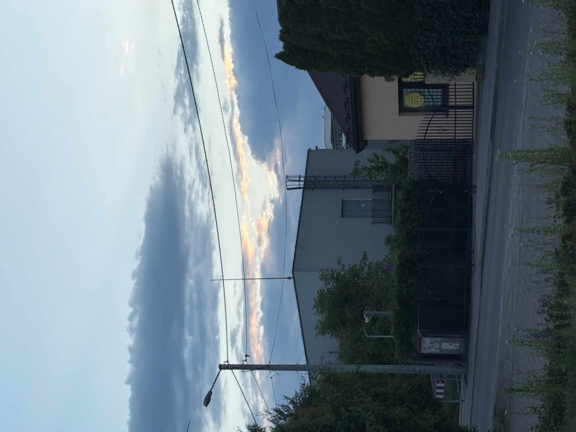 This image depicts a street scene at an unspecified time, likely dusk or dawn, given the light conditions and cloud coloration. The sky features a mix of light blue and dark grey clouds, with a horizontal band of orange and pink hues visible beneath a darker cloud formation in the middle distance. Overhead, multiple utility lines traverse the frame.
In the foreground, a paved road or sidewalk is visible, with a narrow strip of unkempt grass and weeds lining its edge.
The midground is dominated by structures and vegetation. On the left, a tall grey utility pole with a street light extends upwards, supporting multiple power and communication lines. Dense green foliage, including trees and bushes, partially obscures the base of the pole and extends along a fence line. Behind this foliage, a light-colored utility box is visible next to a dark metal fence.
To the right, a peach-colored building with a dark-tiled, sloped roof stands prominently. A window on its side displays a circular yellow sign with black text, partially visible. A tall, dark green evergreen bush or tree is positioned directly in front of this building, obscuring part of its facade. A black ornate metal gate is closed in front of the peach-colored building, with another section of metal fencing to its left.
In the background, behind the street light and the dense foliage, a large, light grey, possibly industrial-style building with a flat roof is present. An external metal ladder or scaffolding structure is attached to its side, leading up to the roof. A doorway with a small balcony is visible on the building's left side. Further in the background, behind the peach-colored building, another structure with a distinctive roof shape can be faintly discerned.
There are no people or animals visible in the scene. The location is in the city of Unknown, Unknown.