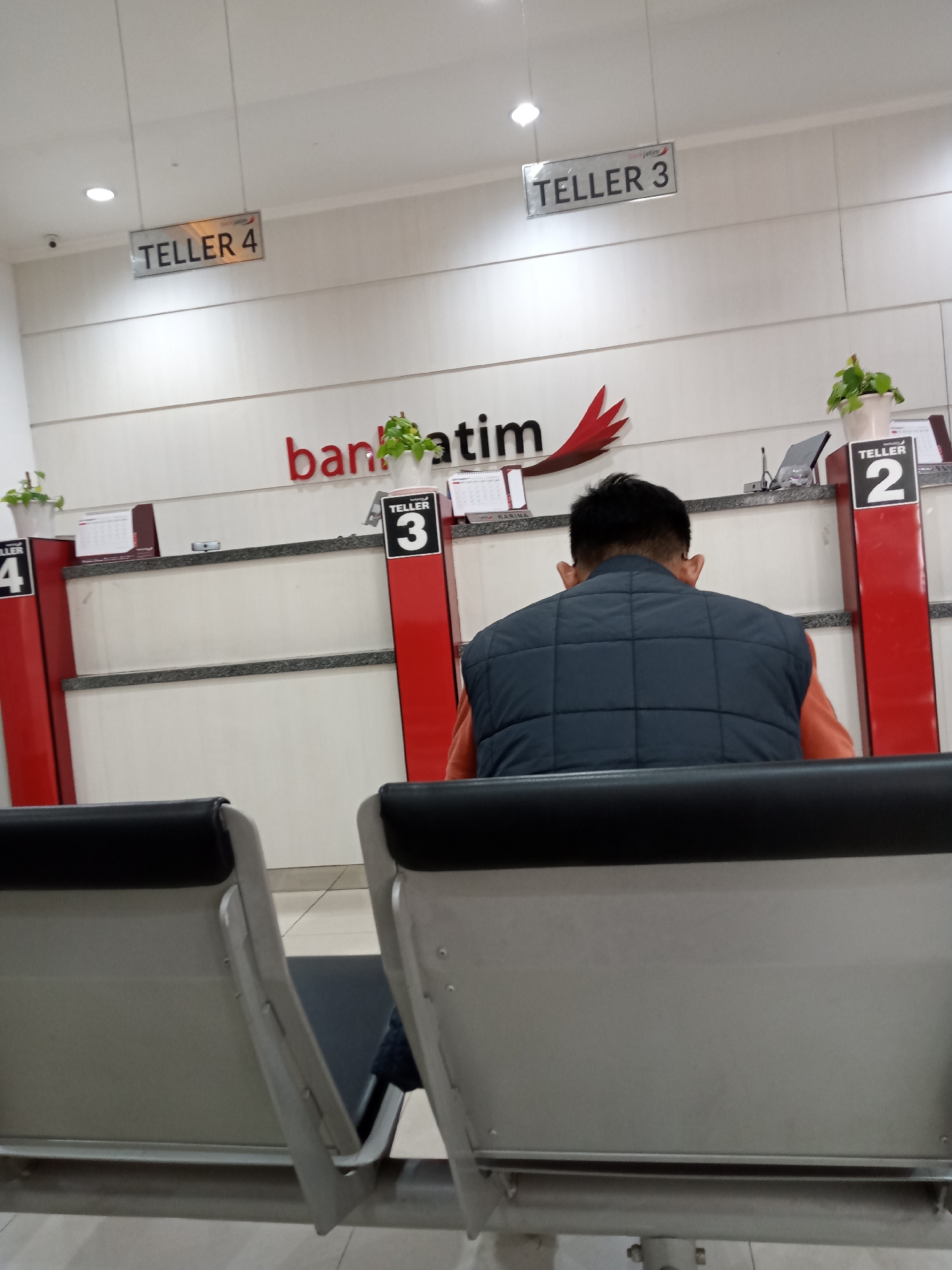 The image depicts the interior of a bank, likely a branch of Bank Jatim, in Batu, Indonesia. In the foreground, two empty waiting chairs are visible, suggesting a queue or a period of low customer traffic. A person is seated in one of the chairs, facing away from the camera, wearing a quilted dark vest over an orange shirt. This individual is positioned in front of a bank counter area. Above the counter, several signs are displayed. A large red and white sign with the text "bank jatim" is prominently featured. To the left, a sign reads "TELLER 4", and to the right, another sign indicates "TELLER 3". Further to the right, a red pillar displays "TELLER 2". The counter itself is equipped with what appear to be transaction areas, and small potted plants are placed on top of the counter, adding a touch of greenery to the institutional setting. Calendars are also visible on the counter, indicating the date. The overall environment appears clean and well-lit, consistent with a professional banking establishment. The specific location is identified as Batu, Indonesia, a city known for its tourism and cooler climate, which might be relevant to the interior design and customer experience of the bank branch.