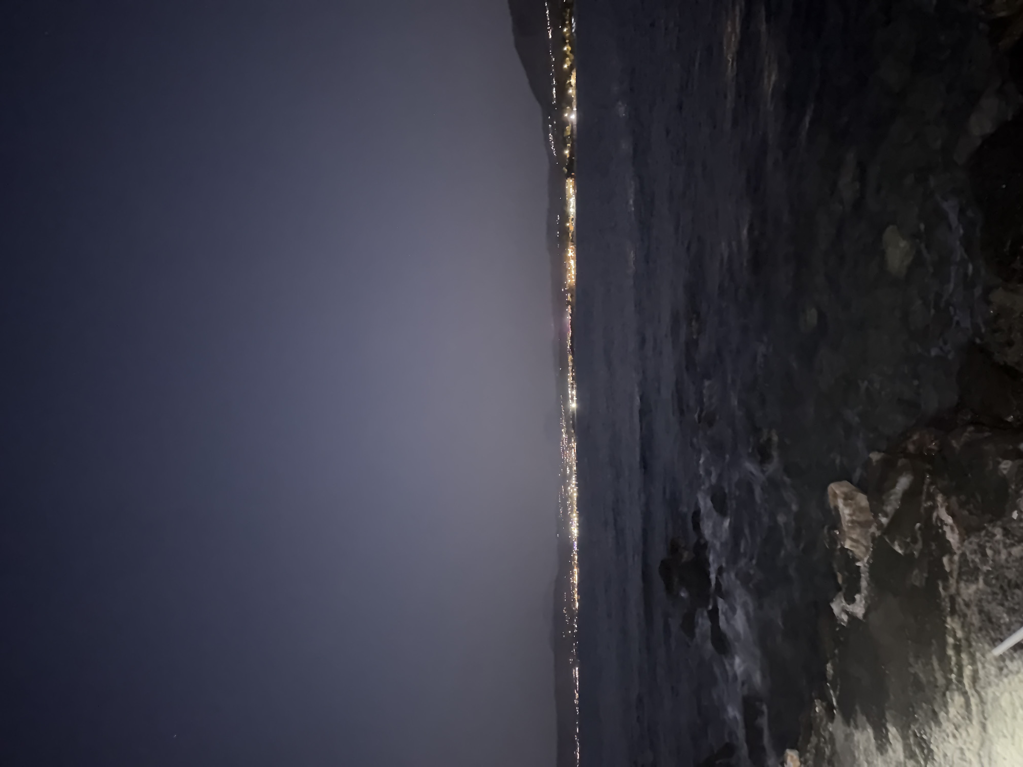 The image depicts a nighttime scene of the coastline near Georgioupoli, Greece. The dark, choppy sea fills the foreground, with waves lapping against a rocky shore. In the background, a distant city skyline is illuminated by a multitude of lights, suggesting a populated area. The sky is a deep, dusky purple, with a faint haze obscuring the horizon. No people or specific landmarks are visible. The overall impression is one of a serene, nocturnal coastal view.