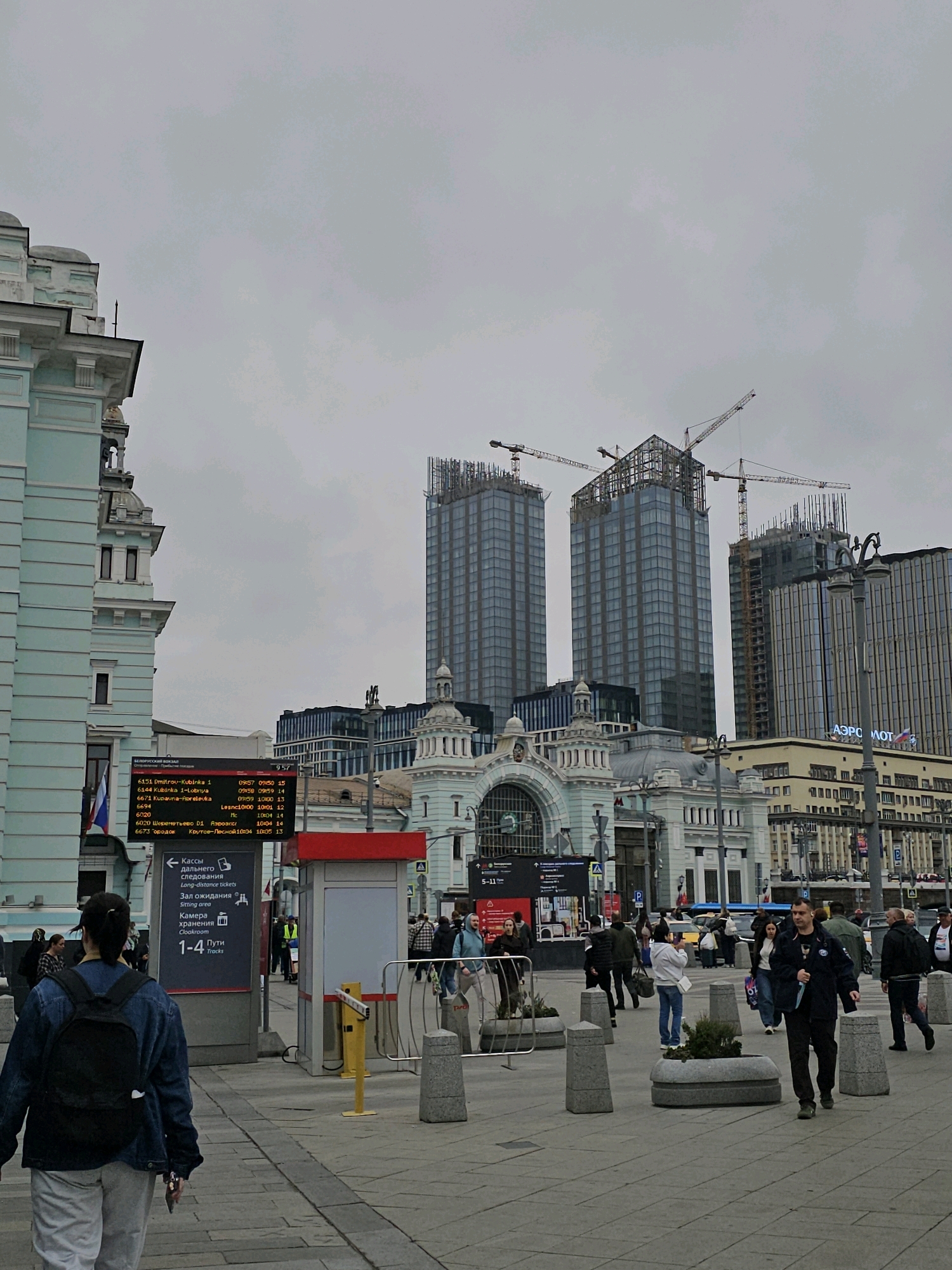 The image captures a street scene in Moscow, Russia, likely near a transportation hub, given the presence of a departure board and signage for "Tracks" and "Waiting Room." In the foreground, a young woman with a backpack walks away from the camera. Several other pedestrians are visible, some walking in various directions, others seemingly waiting or conversing. A yellow barrier is set up on the sidewalk. In the mid-ground, a red and white kiosk stands. Behind this, a large, ornate building, possibly a train station, features arched windows and classical architectural details. Beyond the station, modern skyscrapers are under construction, with cranes visible against an overcast sky. One building prominently displays the "Aeroflot" logo. A digital display board shows train departure information with times and destinations. The overall atmosphere appears to be that of a busy urban environment with a mix of historical and contemporary architecture.