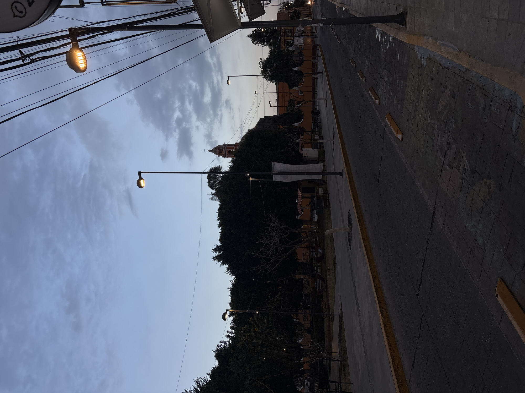 The image depicts a street scene in San Andrés Cholula, Mexico, during twilight hours, characterized by dim ambient light and active street illumination. The sky is a gradient of dark blue to light grey, partially covered by scattered clouds.
In the foreground, a paved street, likely made of stamped concrete with a tiled pattern, runs from the bottom center into the distance. It is bordered by yellow painted curbs on both sides. Along the right side of the road, several light brown rectangular concrete wheel stops are evenly spaced. The sidewalk on the immediate right is also paved.
To the left of the street is a public park or plaza area featuring dense, dark green trees, some with full foliage and others with bare branches. Several park benches are visible along a paved pathway within the park. Multiple tall streetlights with modern, warm-toned LED fixtures illuminate sections of the park and the street.
On the right side of the image, a robust utility pole stands prominently, supporting a dense network of black electrical and communication cables that crisscross the upper right quadrant of the frame. An illuminated street lamp with a bright, warm yellow light is affixed to this pole, facing downwards towards the street. Another street lamp is visible further down the street on the right. A circular sign with a phone icon is attached to the utility pole. A partial view of a building or structure with a white overhang is present on the far right. A single car headlight is faintly visible in the far distance on the right side of the road, suggesting an approaching vehicle.
In the background, a distinct red and orange church tower, topped with a cross, rises above the tree line. This is identifiable as the tower of the Parroquia de San Andrés Apóstol. Below and surrounding the church tower are orange-colored buildings, indicating a structured urban environment. String lights are visible, stretched horizontally across the street leading towards these background structures. No people or overt actions are present in the scene.