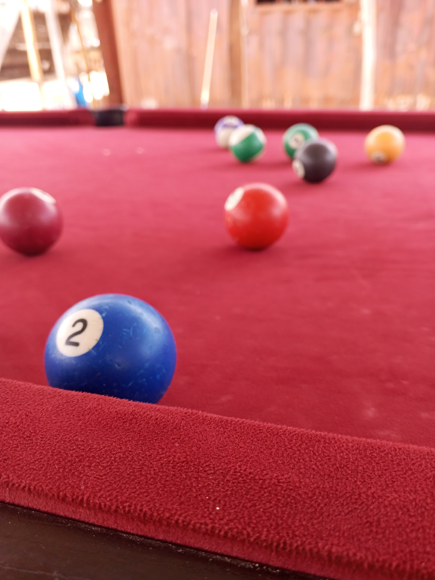 The image shows a close-up, low-angle view of a red felt billiard table with several pool balls scattered on it. In the foreground, a blue ball with the number '2' clearly visible is positioned in sharp focus. To the left and slightly out of focus is a solid red ball. In the mid-ground, several other balls are visible, including a solid red ball, a dark-colored ball with a white stripe, a green ball, and a yellow ball. The background is blurred, showing wooden structures that suggest an outdoor or semi-outdoor setting, possibly a patio or a covered area. The lighting appears to be natural daylight, casting soft shadows. The overall impression is one of a casual game of billiards being played outdoors. There is no visible text beyond the number on the blue ball. The location context of Lokoja, Nigeria, is noted but not directly discernible from the visual elements in the image itself.