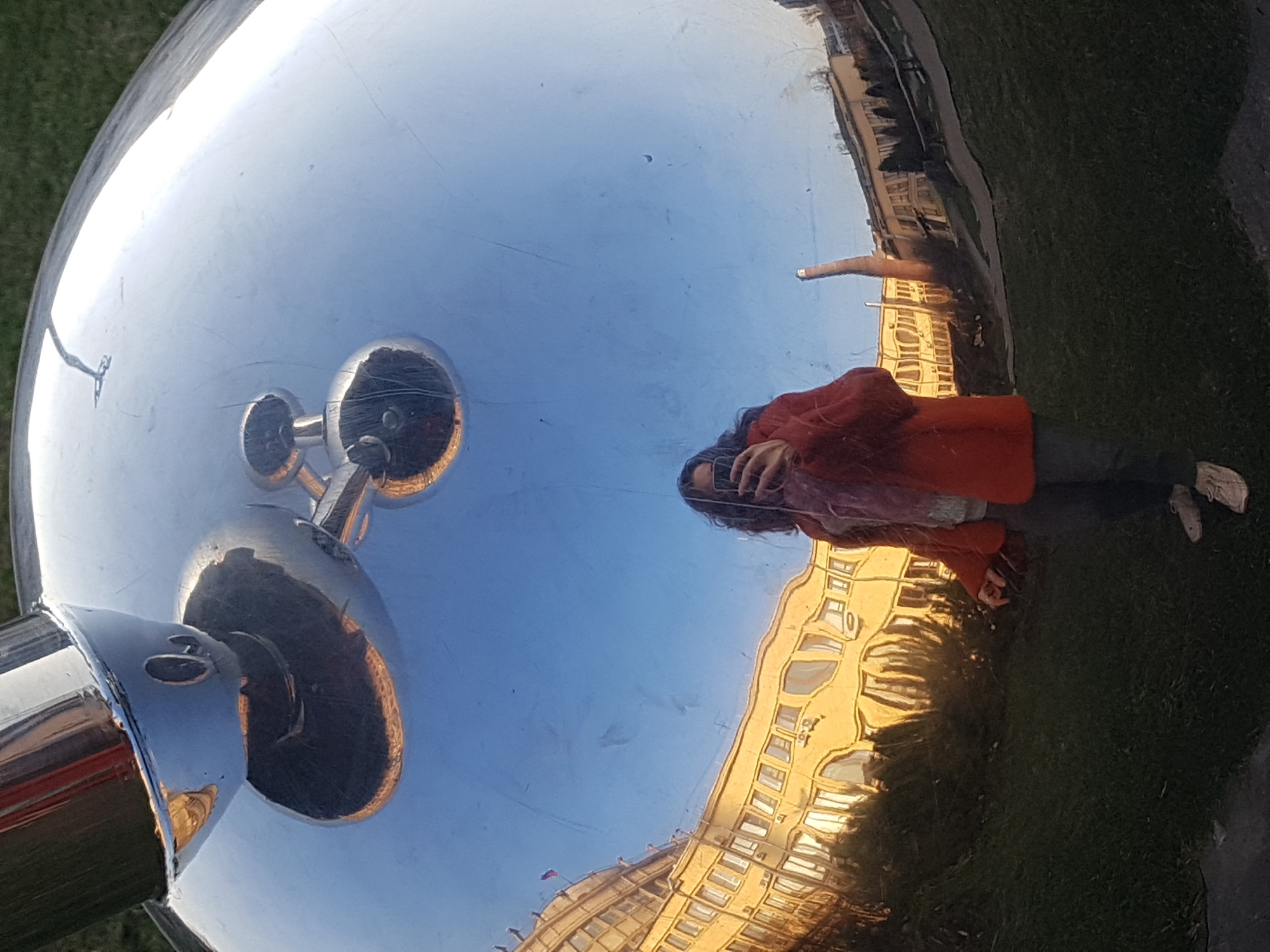 The image captures a distorted reflection in a polished, metallic sphere. The reflection includes a person wearing a red jacket and holding a phone, buildings with numerous windows, a grassy area, and a lamp post with spherical bulbs. The Russian flag is visible above the reflected building. Based on the architectural style of the building, the presence of the flag, and the type of lamp, the location can be identified as Saint Petersburg, Russia.