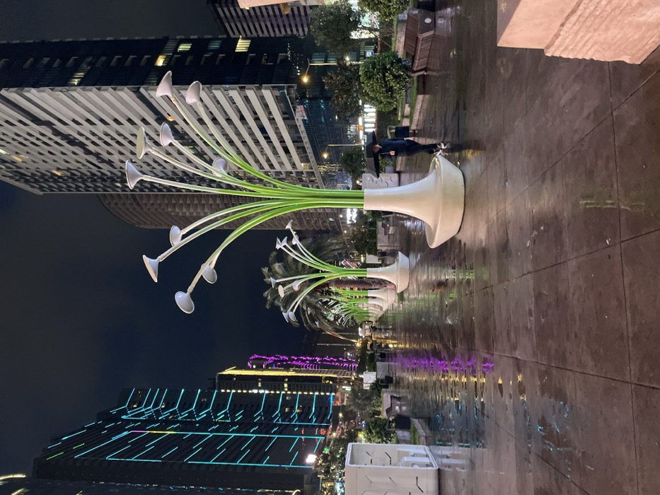 The image depicts a nighttime urban scene on a wet, tiled promenade in Batumi, Georgia. A row of distinctive modern streetlights, stylized as green-stemmed plants with white, bell-shaped luminaires housed in large, white vase-like bases, extends into the background. The ground is reflective from recent rain, showing blurred reflections of neon and artificial lighting.
On the left, a tall, dark high-rise building is illuminated with prominent horizontal and diagonal neon-blue light strips, and another building behind it features vertical purple lighting. On the right, a multi-story building with a light-colored, patterned facade featuring horizontal elements is visible, with another dark building to its right. Several palm trees are positioned behind the stylized streetlights, and additional smaller bushes and planters line the promenade.
A single individual is observed on the right side of the promenade, walking away from the viewer. They are holding a black umbrella open above their head and are dressed in dark clothing with light-colored shoes. The sky above is dark.