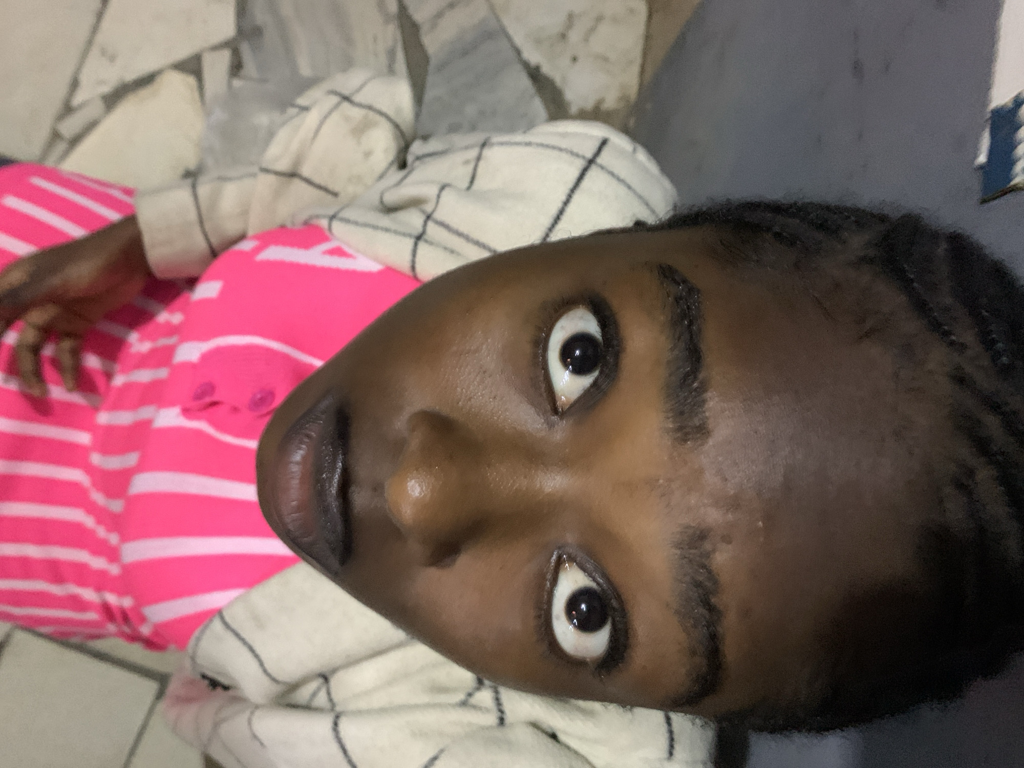 In an unspecified location within New Stadium, Nigeria, the image presents a close-up, upward-facing perspective of a dark-skinned individual, likely a young adult male, positioned on a ground-level surface. The individual's eyes are wide open with significantly dilated pupils, reflecting a direct gaze toward the camera, indicating a state of surprise or intense focus. The nose is broad, and the full, dark lips are slightly parted. Short, dark hair or braids are visible at the lower edge of the frame.
The individual is wearing a vibrant pink garment featuring vertical white stripes, which appears to be a shirt or dress with partial buttons visible on the right chest area. Over this, an off-white or cream-colored piece of clothing, possibly a sweater or blanket with a subtle dark grid pattern, is draped around the neck and shoulders.
The background consists of a light-colored floor, likely composed of tiles or stone, exhibiting visible grout lines and some irregular surface textures. A blurry, small object with blue and white elements is partially visible in the bottom right corner of the frame. The overall lighting appears dim, suggesting indoor or low-light conditions, with reflections indicating a direct light source or flash.
