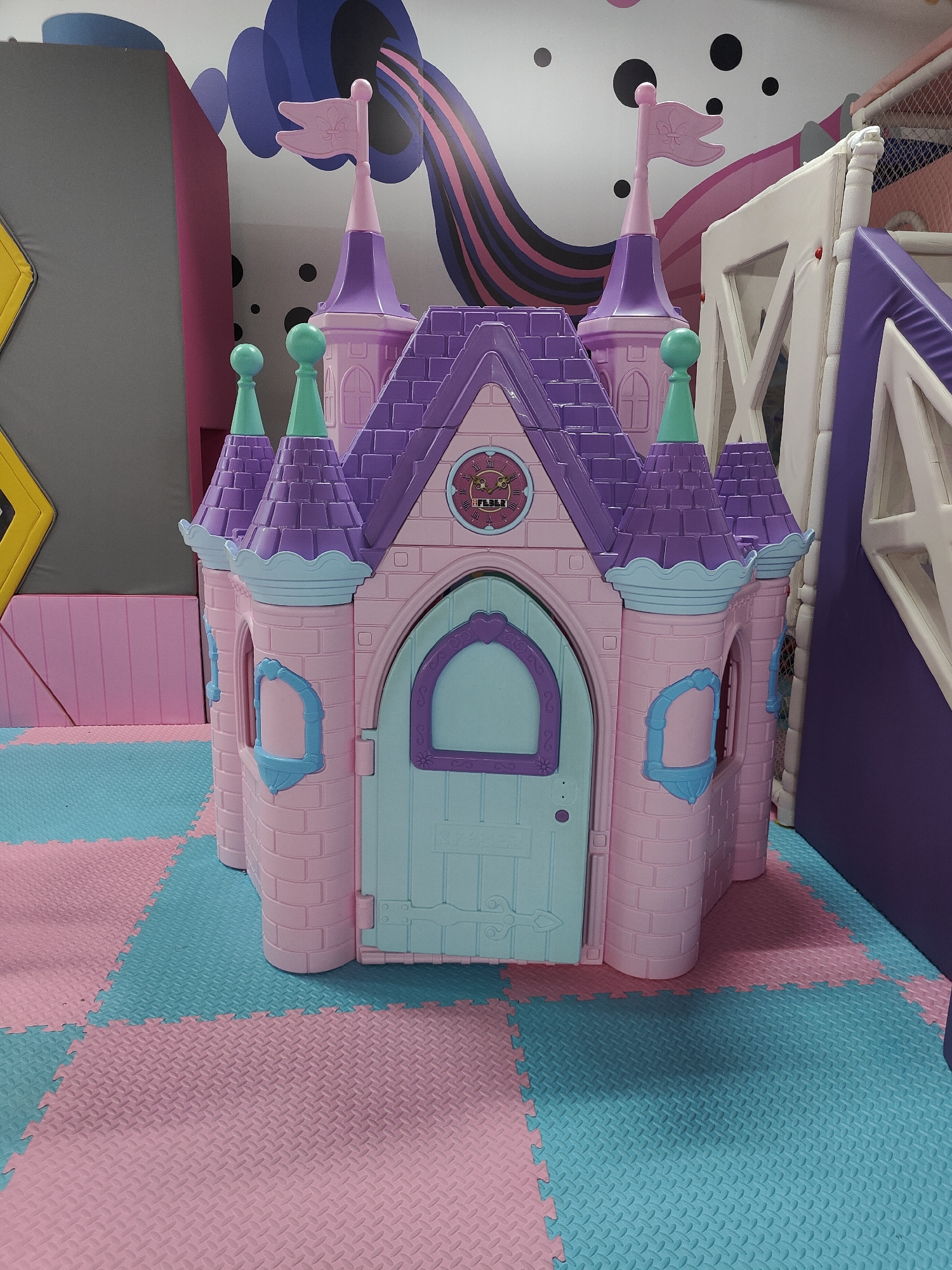 A pink plastic play castle is the main subject in this medium shot. The castle has purple turrets and flags, and a light blue door. It is placed on a floor made of interlocking foam mats in alternating pink and turquoise colors. To the left of the castle, a large grey padded structure with a yellow and black warning sign is visible. In the background, a mural with colorful abstract shapes and black circles adorns the wall. To the right, a white and purple play structure with a slide is partially visible. The lighting suggests it's daytime indoors. The setting appears to be an indoor playground or play area, possibly for children. No people are visible in the image. The clock on the castle is decorated with roman numerals and appears to be set to approximately 3:30. There is no other discernible text in the image.