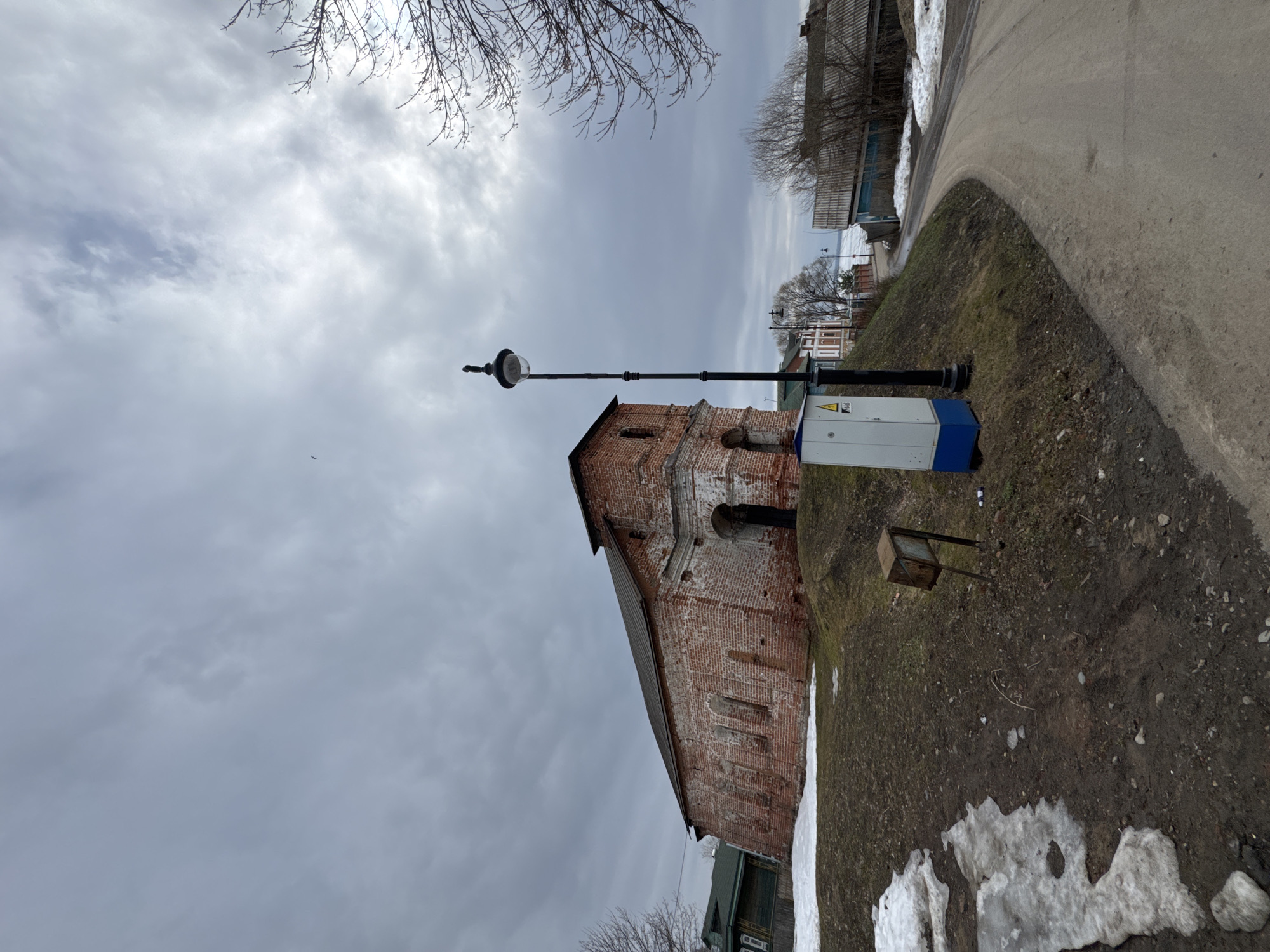The photograph captures an outdoor scene in Ново-Никольское, Russia, likely during late winter or early spring. An aged, brick building with a sloping roof is situated at the left of the frame. A slender, black lamppost extends upwards, partially obscuring the building. Adjacent to the lamppost, and closer to the viewer, is a utility box with a white and blue exterior, bearing a yellow warning sign. The ground is patchy with exposed dirt, grass, and melting snow. A dirt path borders the right side of the image. The sky is overcast with thick cloud cover, creating a diffused, grey light. Bare tree branches appear at the top of the frame.