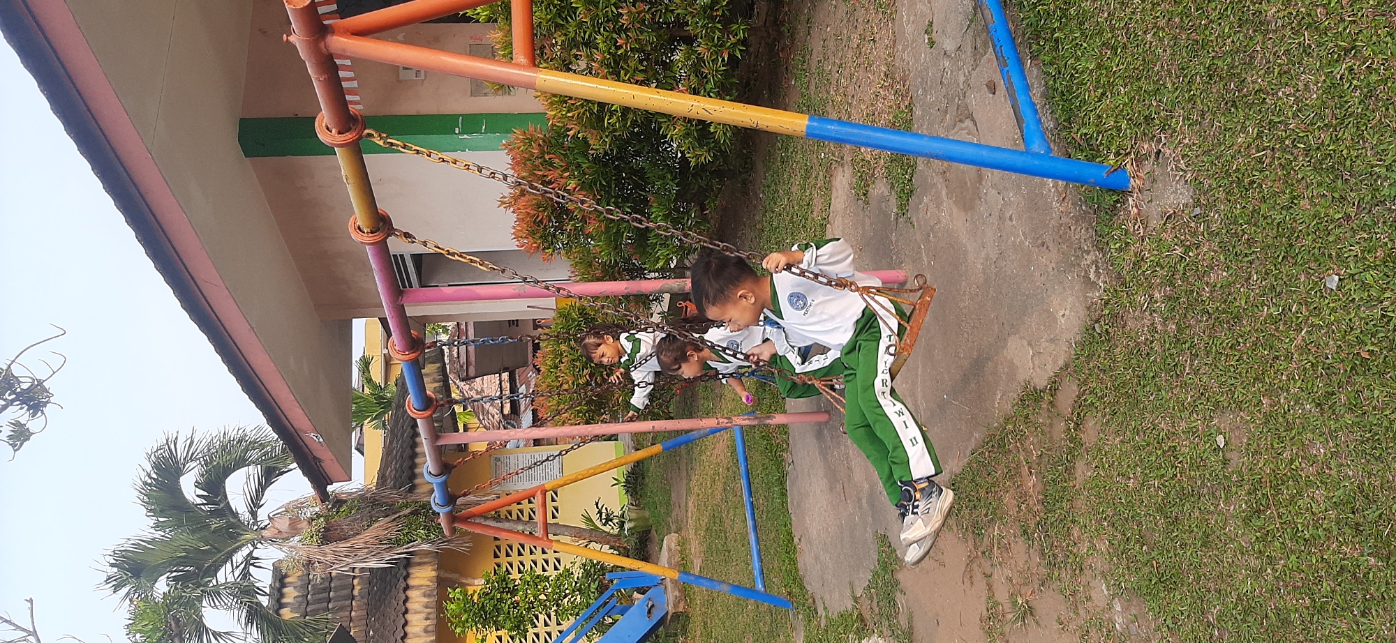In Jambi, Indonesia, three young children, two boys and one girl, are depicted on a playground swing set. The children are wearing matching white long-sleeved shirts with green trim and green pants, suggesting they are in school uniforms. The foremost child, a boy, is seated on a rusty swing, his legs extended forward, his body leaning back, and his arms gripping the chains. He appears to be smiling. Behind him, another child, likely a girl, is also on a swing, reaching out with one hand. A third child, a girl, is visible further back on another swing. The swing set is constructed with brightly colored poles in orange, yellow, pink, and blue. The background shows a building with a reddish-brown roof and yellow walls, with some landscaping including bushes and a palm tree. The ground is a mix of grass and concrete.