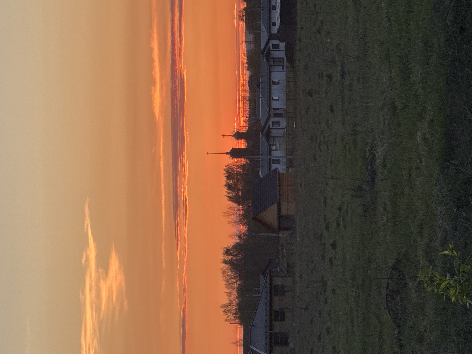 A landscape shows an orange sunset on the left, transitioning to a grassy area with houses on the right. The tops of buildings and a church spire silhouette against the sunset. The location is Горный, Russia.