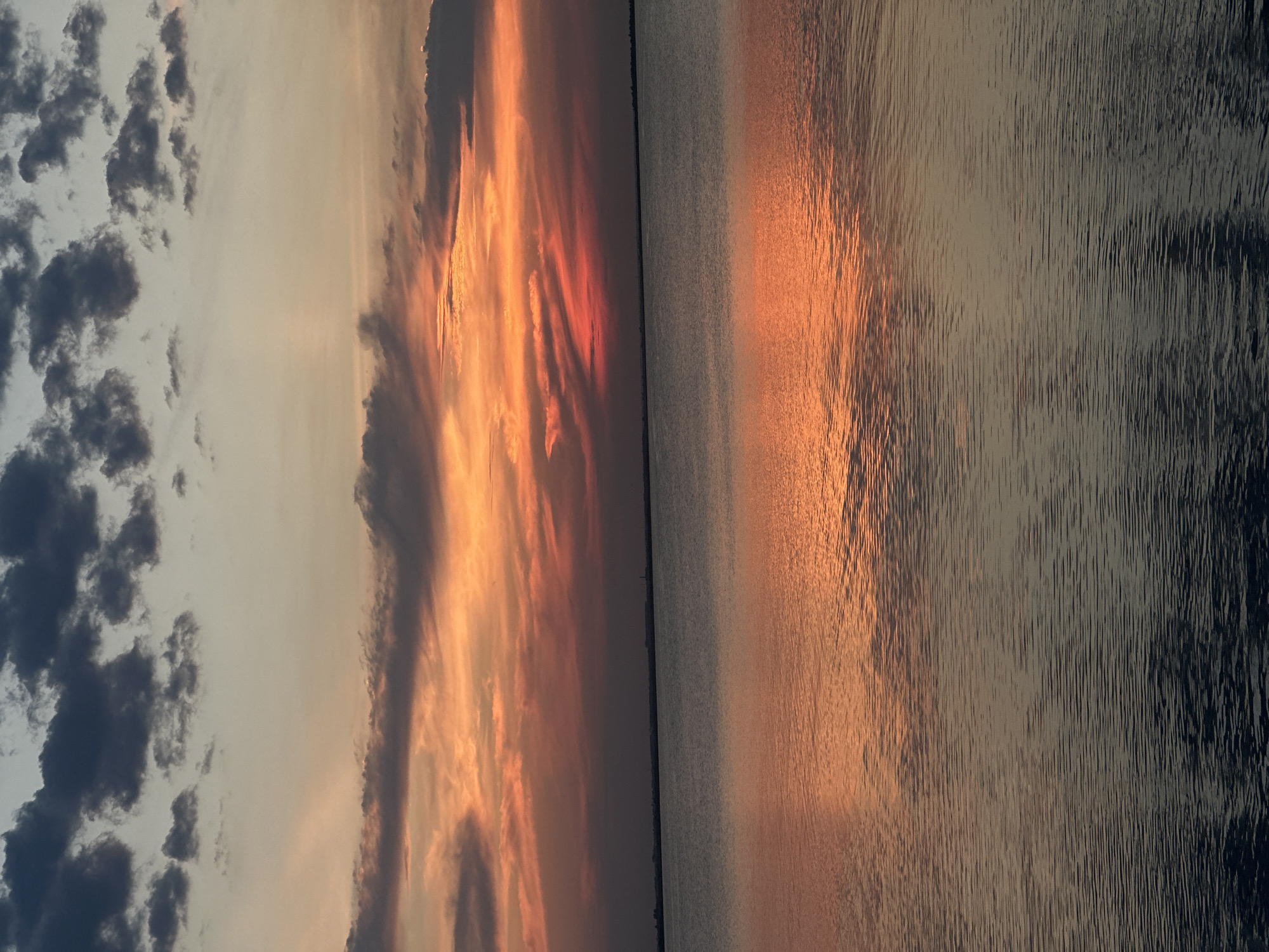 The image depicts a serene sunset over a body of water, likely a lake, given its expanse and the distant, faint landmass on the horizon. The sky is a dramatic canvas of colors, transitioning from deep gray clouds at the top to vibrant oranges and reds closer to the horizon, where the sun is setting. Rays of light emanate from the setting sun, breaking through the clouds and casting a warm glow. The water's surface is calm, with gentle ripples reflecting the fiery hues of the sunset. The reflection is most prominent in the upper portion of the water, mirroring the sky's colors, and gradually deepens to a darker tone as it recedes towards the bottom of the frame. The distant horizon shows a faint outline of land, possibly a city or natural elevation, with a solitary, tall structure, possibly a tower or lighthouse, visible against the sky. The overall atmosphere is tranquil and picturesque. The location is identified as Kokshetau, Kazakhstan, suggesting this is a view of one of its lakes, such as Lake Borovoe or Lake Shchuchye, popular tourist destinations known for their natural beauty, especially during sunset. There are no people or specific objects or actions depicted in the scene, making it a purely environmental landscape.