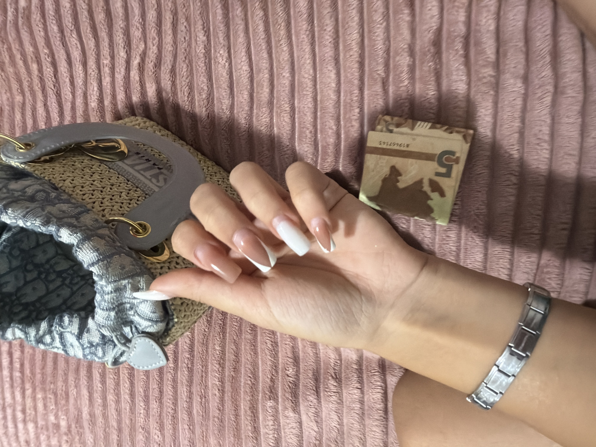 A person's left hand, adorned with French manicure acrylic nails and a silver charm bracelet, rests on a pink, textured fabric surface. Next to the hand, to the right, is a small wad of what appears to be money, with the numeral "5" visible on one of the bills, and a serial number. In the upper portion of the frame, a portion of a woven handbag with grey trim and gold hardware is visible. The scene is devoid of any information or context that would place it in the city of Digah, Azerbaijan.