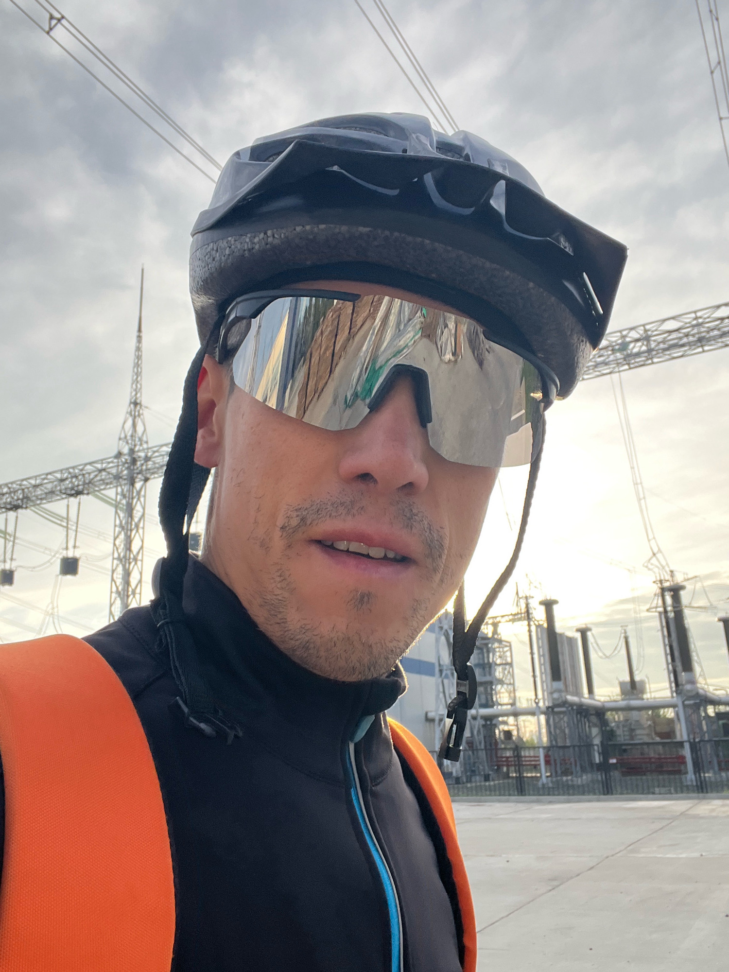 A man wearing a black cycling helmet and silver mirrored sunglasses is looking at the camera. He is wearing a black cycling jacket with an orange strap across his chest, possibly from a backpack. In the background, there is an electrical substation with high-voltage power lines and large industrial structures. The sky is cloudy, and the sun appears to be setting, casting a warm glow. The location is the Chaikovsky Thermal Power Plant in Chaikovsky, Russia.