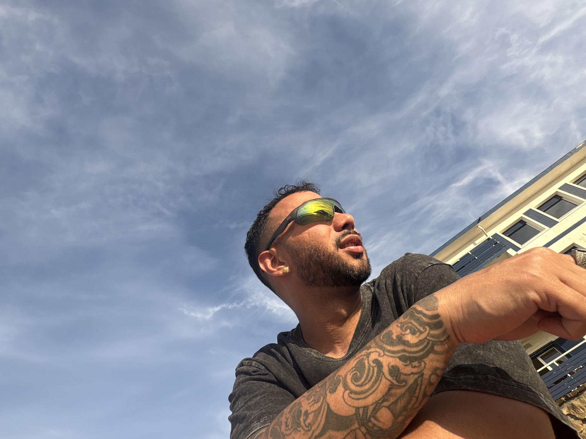 A low-angle, full shot captures a man with tattoos on his left arm, wearing a dark t-shirt and sunglasses. He is positioned in the foreground, with the sky and a building in the background. The sky is partly cloudy with wispy white clouds against a blue expanse. The building appears to be light yellow with blue shutters on the windows. The man's facial features are visible, including his beard and ear jewelry. His left arm, adorned with intricate tattoos, is resting in front of him. The lighting suggests it is daytime, with sunlight casting shadows and reflections on his sunglasses. The scene is set outdoors.