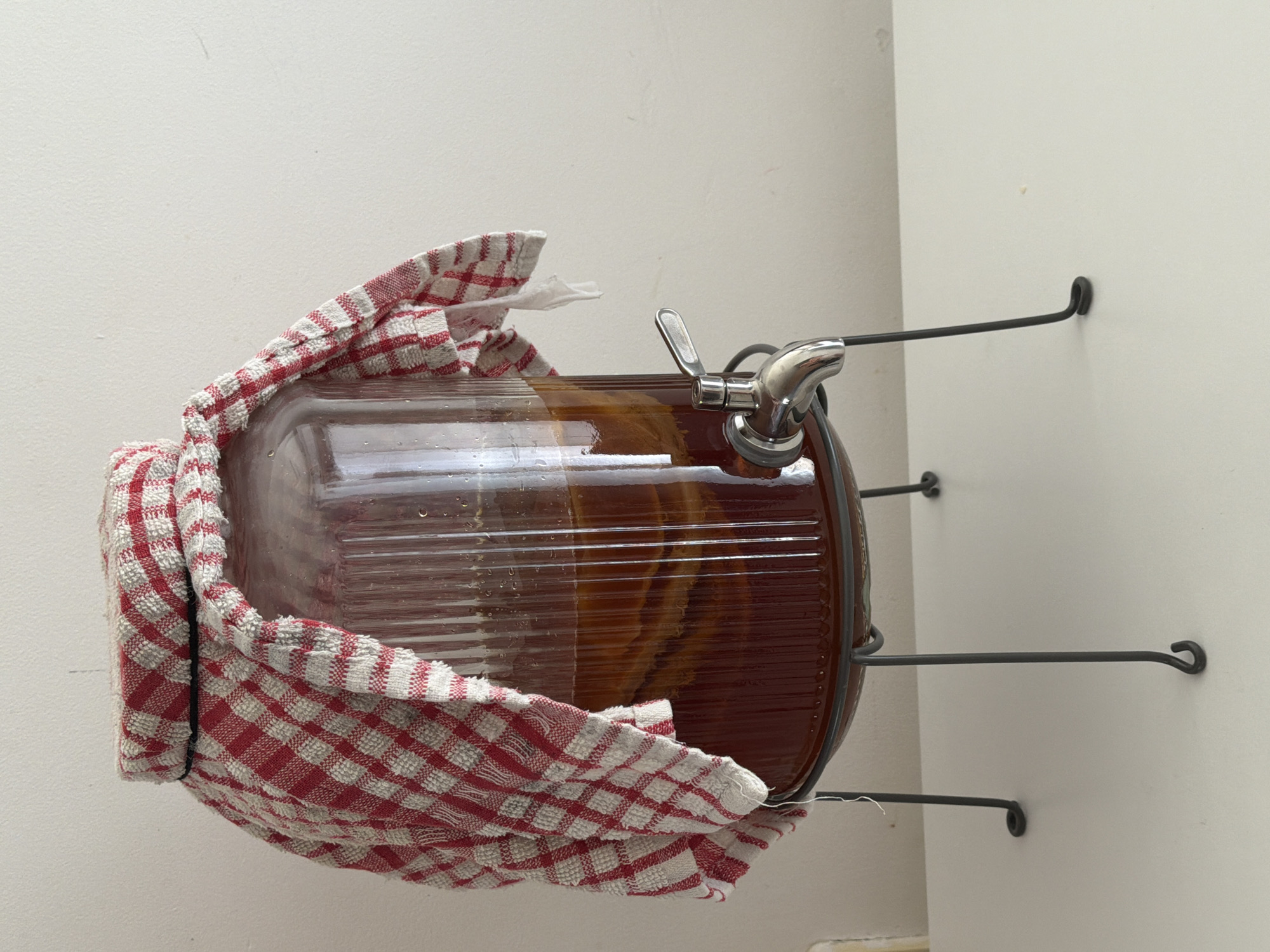 A large, clear glass beverage dispenser, appearing rotated 90 degrees clockwise from its intended vertical orientation, is prominently displayed. It is mounted on a plain, light-colored wall via three black metal hooks that support its cylindrical body. The dispenser is filled with a substantial quantity of dark amber-brown liquid, which exhibits visible layers and textures, characteristic of a fermented beverage such as kombucha. A red and white checkered kitchen towel is draped around the upper portion of the dispenser, potentially for insulation or to cover an opening. A polished metallic spigot with a lever handle is attached near the base of the dispenser (on the right side in the rotated image), indicating its use for dispensing. The wall surface is clean with a slight texture. The scene suggests an indoor setting, likely a home kitchen or pantry. No people or visible text are present in the image. The given location context is Tel-Aviv, Israel.