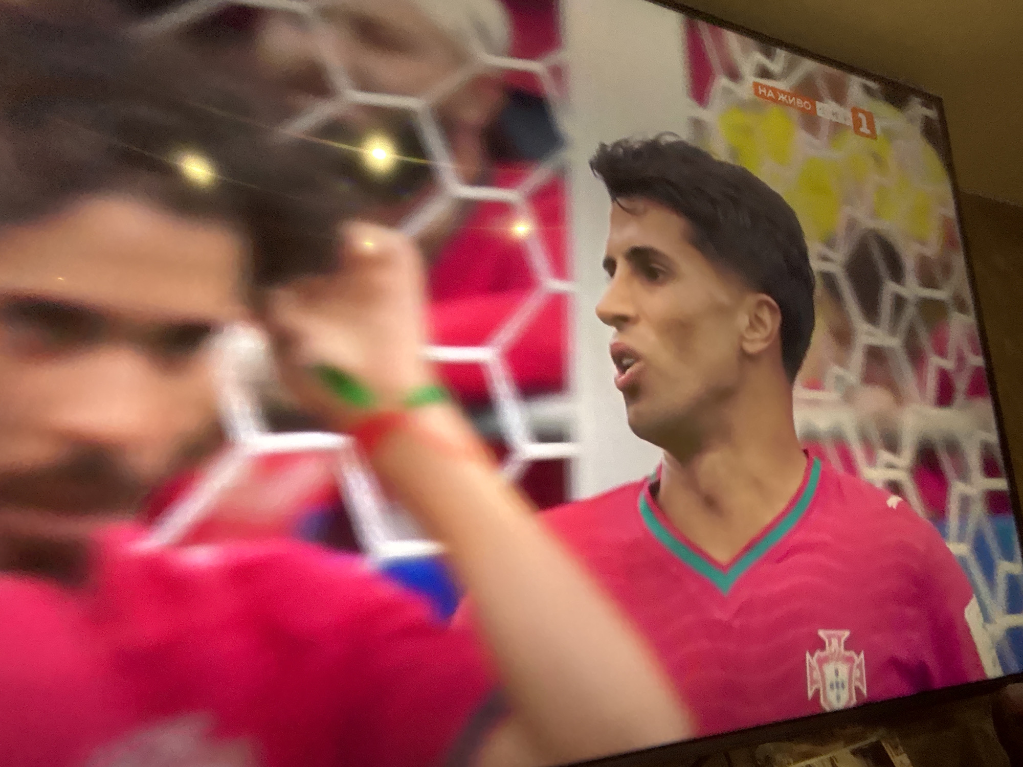 The image shows a television screen displaying a soccer match. On the right side of the screen is a close-up of a male soccer player wearing a pink jersey with a turquoise trim and the Portuguese national team crest. He has dark hair and appears to be speaking. On the left side of the screen, another player's reflection is visible, with a hand in the foreground wearing a red and green wristband. The background on the screen shows a soccer goal net and what appears to be a stadium crowd. In the upper right corner of the screen, there is text that reads "НА ЖИВО" which translates to "LIVE" in Bulgarian, along with a channel logo featuring the number "1". The overall lighting suggests the event is taking place indoors or at night. The presence of the "НА ЖИВО" text indicates the broadcast is live from Bulgaria.