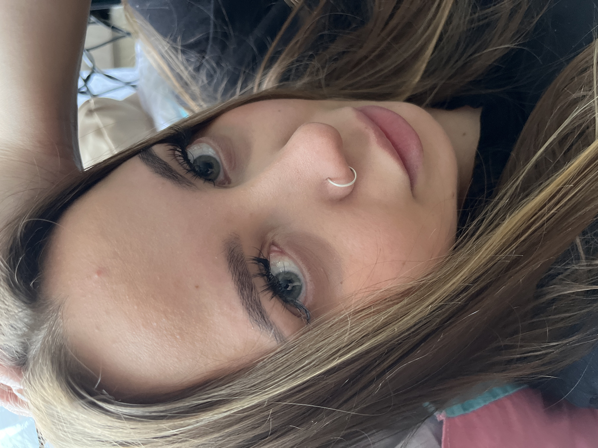 The image shows a close-up, angled portrait of a young woman. She has fair skin, light brown hair with blonde highlights, and light-colored eyes. She is wearing a silver hoop nose piercing. Her eyebrows are dark and well-defined, and she has long eyelashes. The background is blurred and indistinct, suggesting a shallow depth of field. The setting cannot be determined, and the location in Novopokrovskaya, Russia is not identifiable from the image.