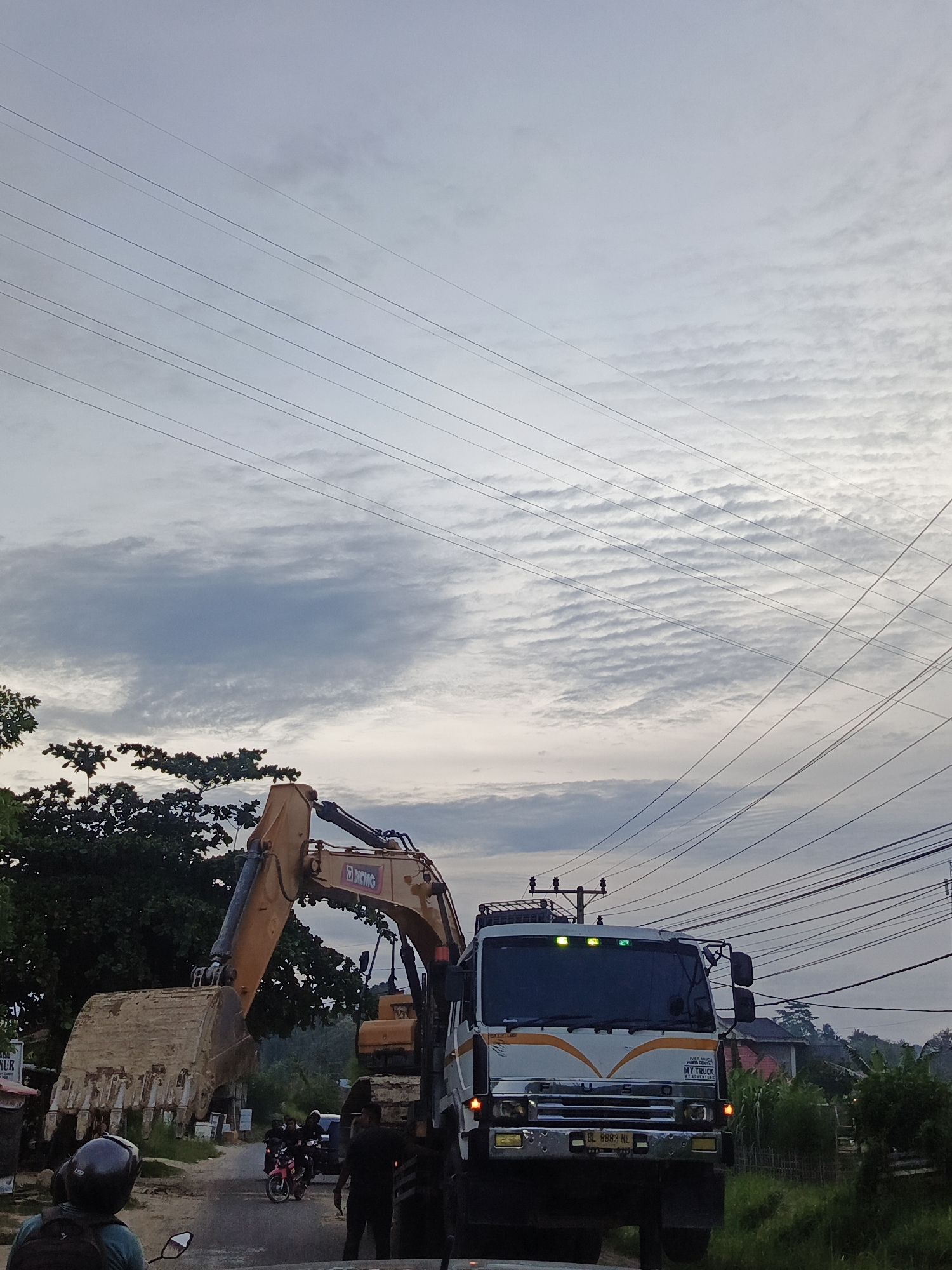 The image shows a construction scene in Lhokseumawe, Indonesia. An orange XCMG excavator is positioned with its bucket elevated, likely being loaded onto a white FUSO truck. Several individuals are standing near the truck. Other motorcycles and vehicles can be seen in the street. Overhead, there are many power lines spanning the scene against a cloudy sky. Vegetation lines the edges of the road.