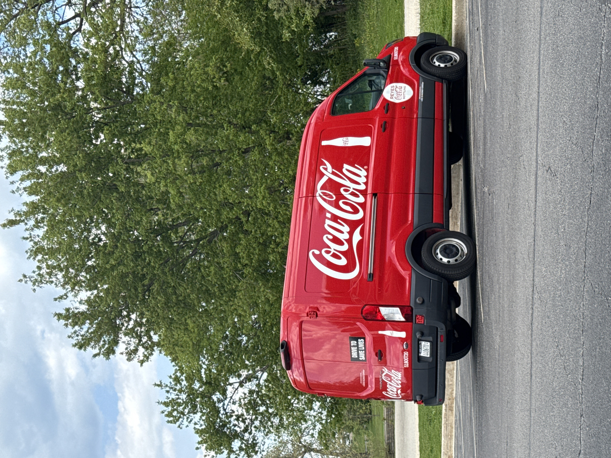 The image shows a red Coca-Cola delivery van parked in a lot, likely for deliveries. The van has the Coca-Cola logo prominently displayed on the side, with a white bottle illustration. The phrase "DRIVE TO SAVE LIVES" is visible on the rear doors. The license plate number is partially visible: 440670. The side of the van also displays the words "REYES Coca-Cola BOTTLING". The setting appears to be a parking lot adjacent to greenery with a view of the sky in the background. The image does not provide any information to place the image in a specific location in Chicago.