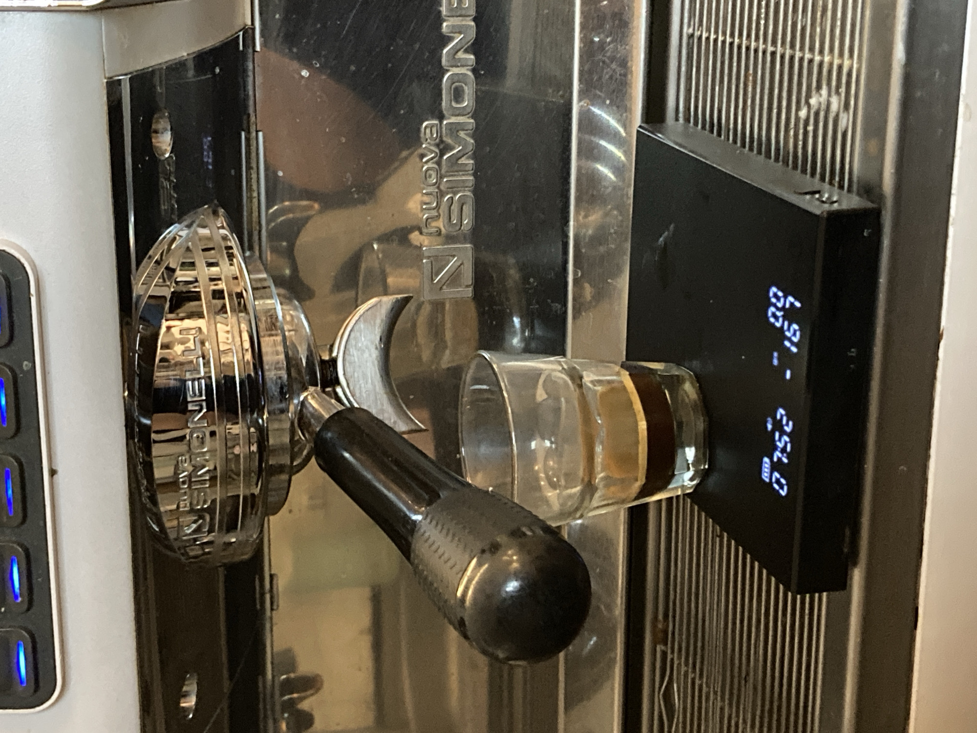 A close-up, rotated shot captures an espresso machine in action. The central subject is a chrome portafilter, reflecting light, with a black, textured handle extending towards the viewer. Below the portafilter's spouts, a clear glass espresso shot glass is positioned, containing a dark brown espresso shot.
To the right, a black digital scale/timer is magnetically or adhesively attached to the side of the espresso machine, positioned in line with the shot glass. Its bright blue LED display is clearly visible, showing "0.0" (likely grams for weight) as the primary large reading, and "0:52.0" (likely minutes and seconds for time) as a smaller reading below it. A small battery icon is also present. The discrepancy between the "0.0" weight reading and the visible coffee in the glass suggests the scale may have just tared, reset after a finished shot, or is not currently weighing.
The espresso machine itself is made of reflective chrome or stainless steel, with the embossed "nuova SIMONELLI" logo prominently displayed on its front panel. To the far left, a section of the machine's control panel is visible, featuring several blue-illuminated rectangular buttons. Behind the scale, a metal grate or cooling fin structure is visible, showing some minor residue or dirt. The overall scene suggests a professional or high-end home espresso setup, with bright, possibly artificial, lighting reflecting off the machine's surfaces.