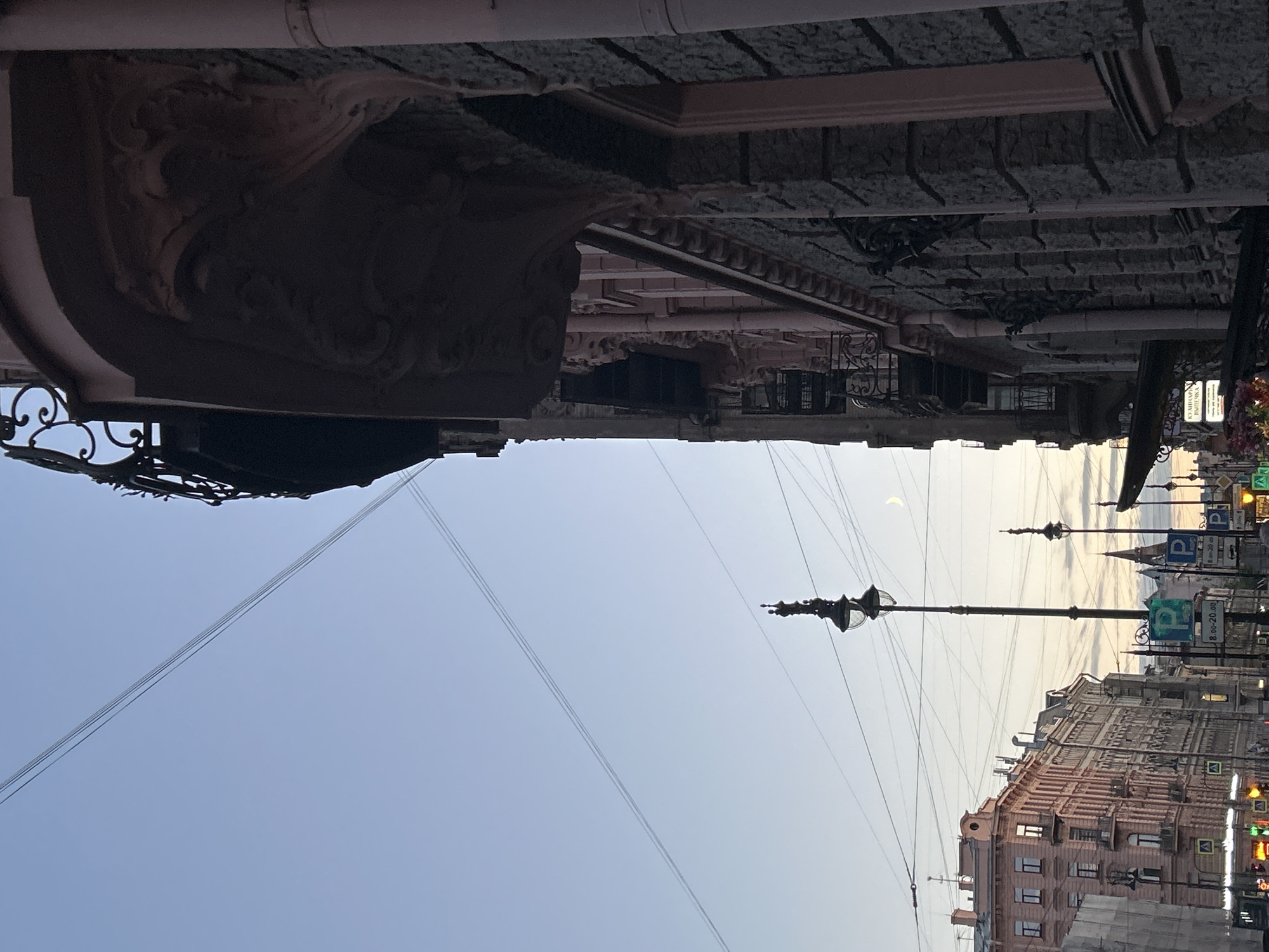 The image depicts a street scene in an urban environment, likely at dusk or dawn, given the soft, pale sky and the crescent moon visible. On the right side of the frame, a detailed architectural facade of a building is prominently featured, characterized by ornate carvings, a balcony with intricate metalwork, and textured stone cladding. To the left, a row of multistory buildings with a pastel-pink facade lines the street, displaying numerous windows and architectural details typical of European cityscapes. The street itself is equipped with lampposts, traffic signals, and various street signs, including parking signs with the letter 'P' and times indicating restricted parking zones. Overhead, a network of wires crisscrosses the sky, suggesting the presence of utility infrastructure. The overall atmosphere is one of quiet urban life, with no visible people or vehicles in the immediate foreground, allowing the architecture and the atmospheric lighting to dominate the scene.