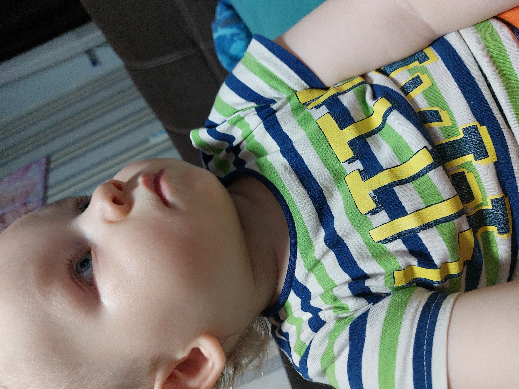The image shows a close-up of a baby or toddler lying on their back. The child is wearing a striped shirt with blue, green, and white stripes, and yellow graphic designs. The child's face is turned towards the left, with fair skin, blue eyes, and light-colored hair. The background includes a hint of a gray, upholstered surface and a light blue or teal-colored object. The location is unknown, and is not possible to infer from the image if it was taken in Chaikovsky, Russia.