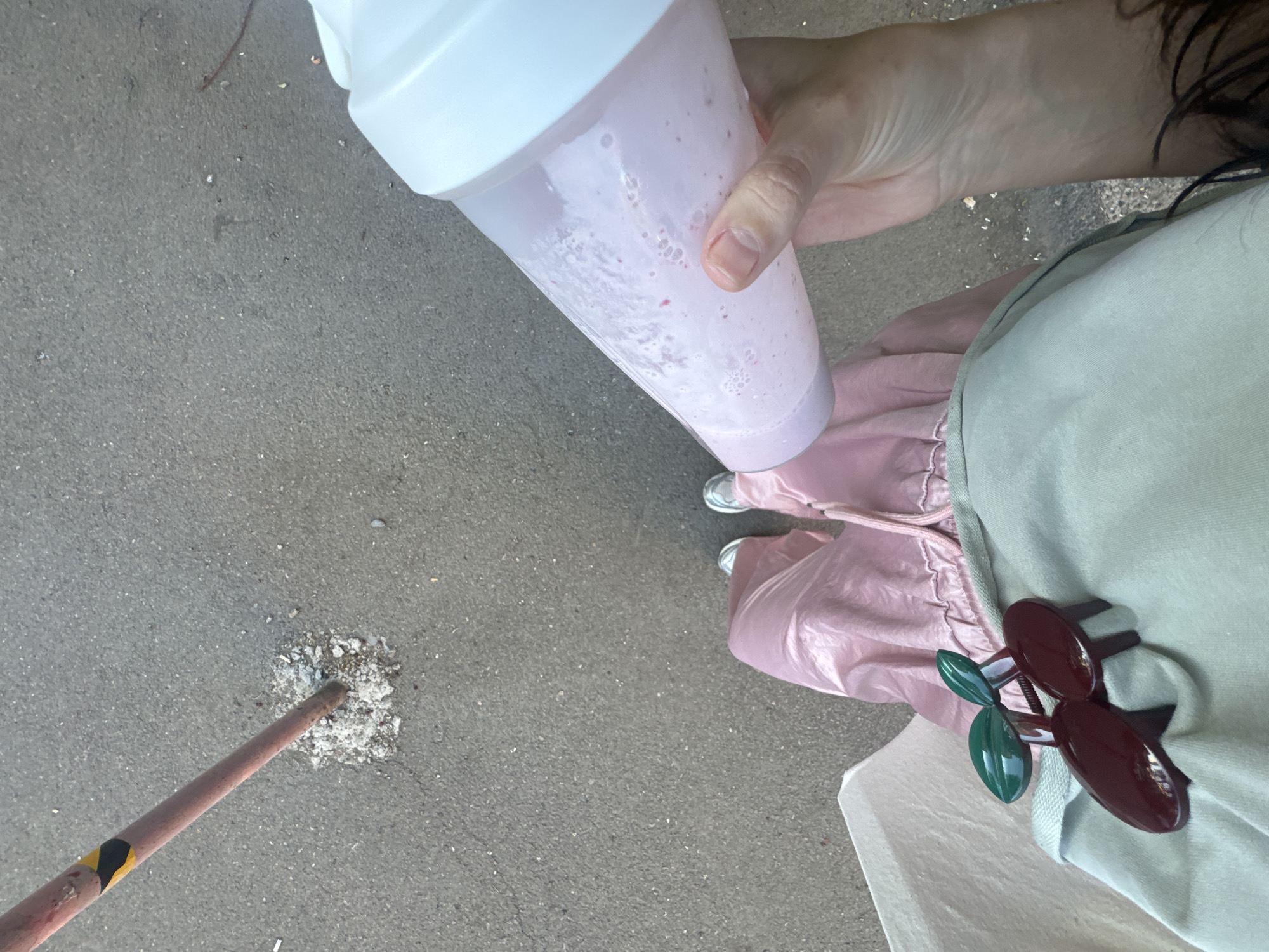 An overhead shot captures a person standing on asphalt or concrete pavement in Krasnoyarsk, Russia. The individual is partially visible, showing their lower torso, legs, and one hand. They are holding a clear shaker bottle with a white lid, containing a frothy pink beverage, likely a smoothie or protein shake, with visible small fruit pieces or bubbles.
The person is dressed in light pink, possibly track pants or a voluminous skirt with a drawstring, and wears light-colored sneakers with white soles. A light green or beige top, possibly a hoodie or apron, is visible around their mid-section. Attached to this garment is a distinctive dark red and green hair clip, shaped like a pair of cherries.
In the foreground and to the left, a reddish-brown metal pole with a yellow and black striped warning band emerges from the ground. It is anchored in a patch of broken concrete or gravel within the pavement. The overall lighting suggests it is daytime. The scene depicts an ordinary outdoor moment in an urban or semi-urban setting.