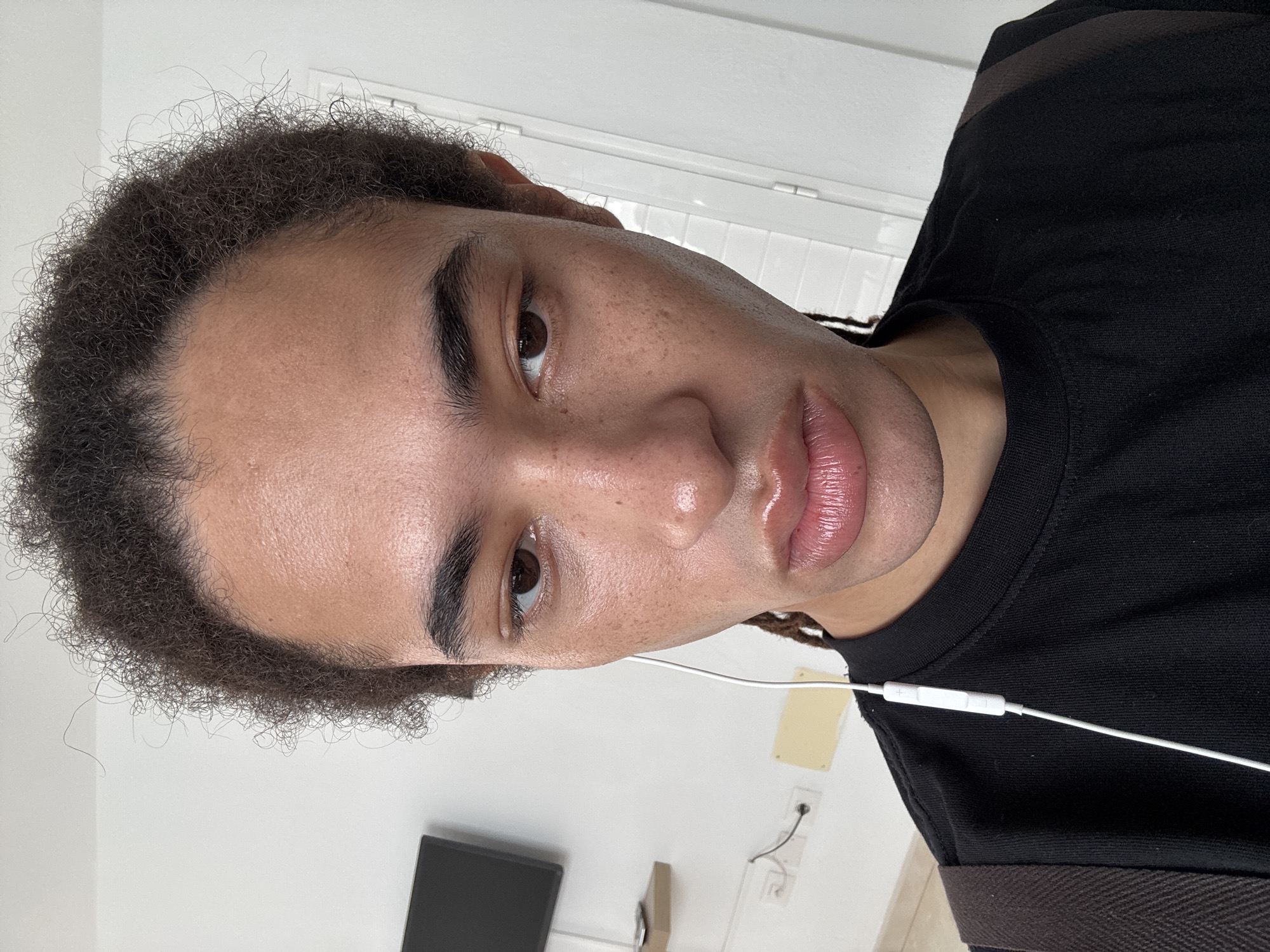 A close-up, slightly angled selfie shows a person with brown eyes and dark, tightly curled hair that appears to be in a natural state. The person has smooth skin with visible freckles and a full lower lip. They are wearing a black T-shirt, and a white earbud cable is visible near their neck. In the blurred background, a white wall with a small, dark monitor and a light-colored electrical outlet are seen. The location cannot be determined with available information.