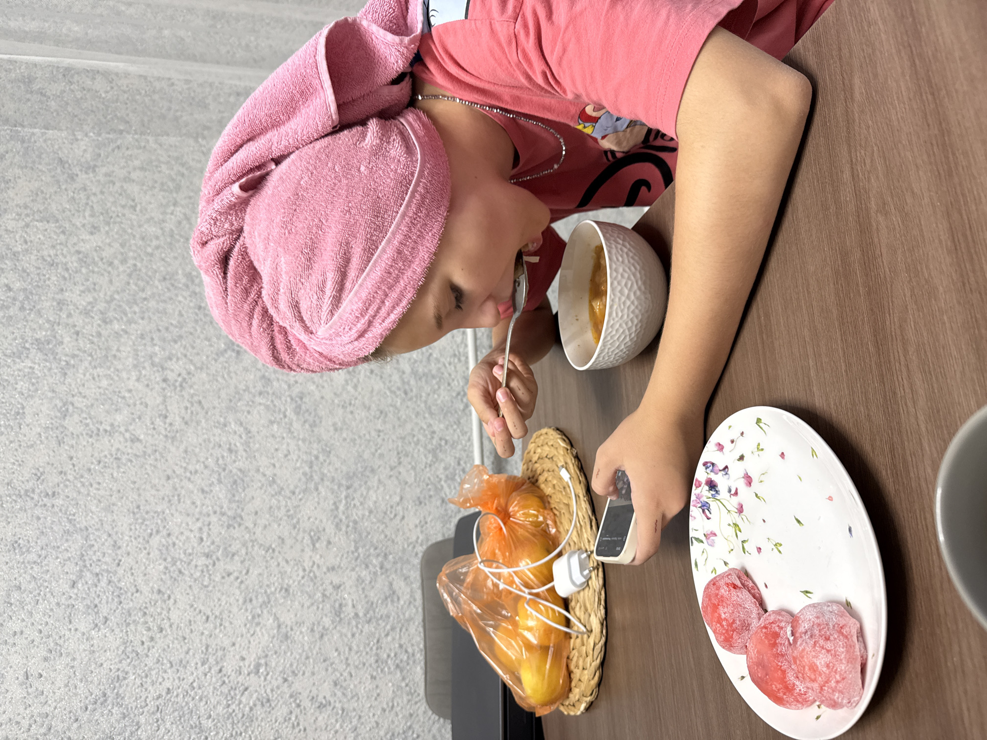 The image shows a young person with a pink towel wrapped around their head, wearing a pink t-shirt, sitting at a wooden table. The person is in the process of eating something from a white bowl with a spoon. On the table, there is also a white plate with three pink, powdery snacks and a small woven basket containing a plastic bag of yellow fruits, a phone, and a white power adapter and cable. The background shows a textured grey wall on the left and the wooden table surface. The scene appears to be indoors, likely in a kitchen or dining area. The lighting suggests it is daytime. The person's facial expression is focused on eating.
