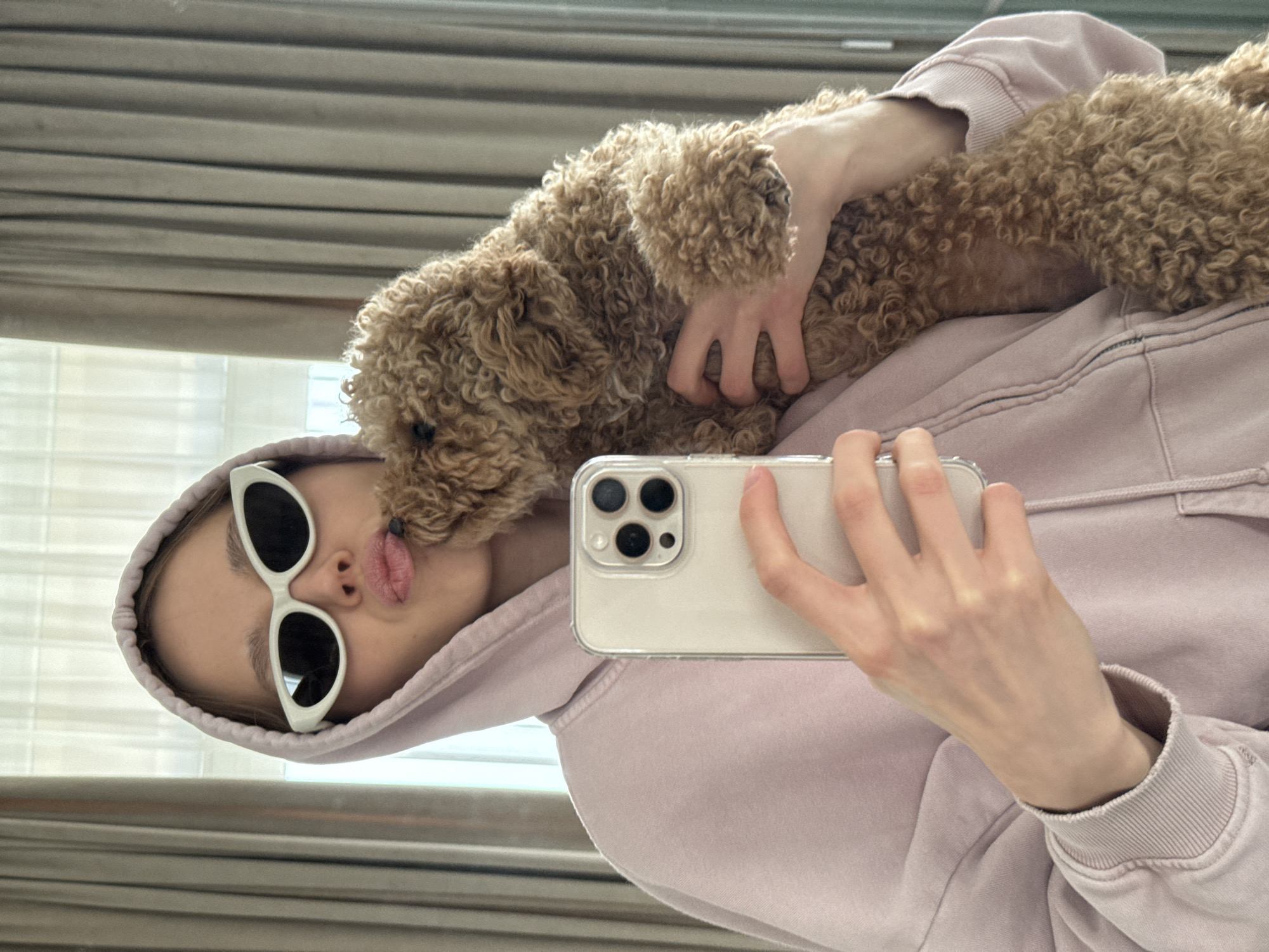 A woman wearing white-rimmed sunglasses and a light purple hoodie is taking a selfie with a brown poodle in an indoor setting. The woman is holding a smartphone, and the dog is resting on her chest. The background includes a window with blinds.