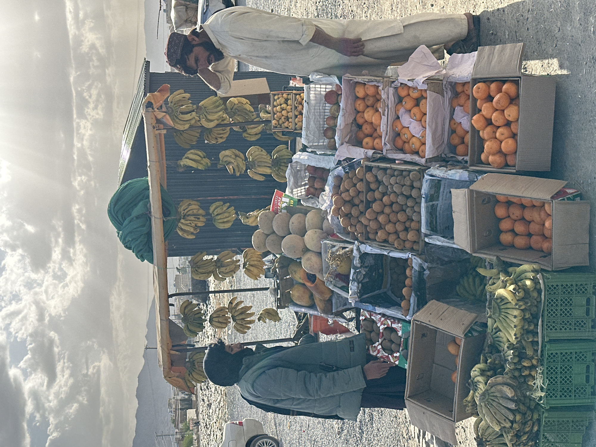The image depicts a roadside fruit stand in Quetta City Tehsil, Pakistan. Two men are present; one, dressed in a white garment and a patterned cap, rests an elbow on the fruit stall. The other man, wearing a dark coat and hat, stands near the front of the stall. The stall is stocked with various fruits, including bananas, oranges, melons, apples, and kiwi, arranged in boxes and crates. The background shows a cityscape and mountains under a cloudy sky. A white car is parked near the stand.