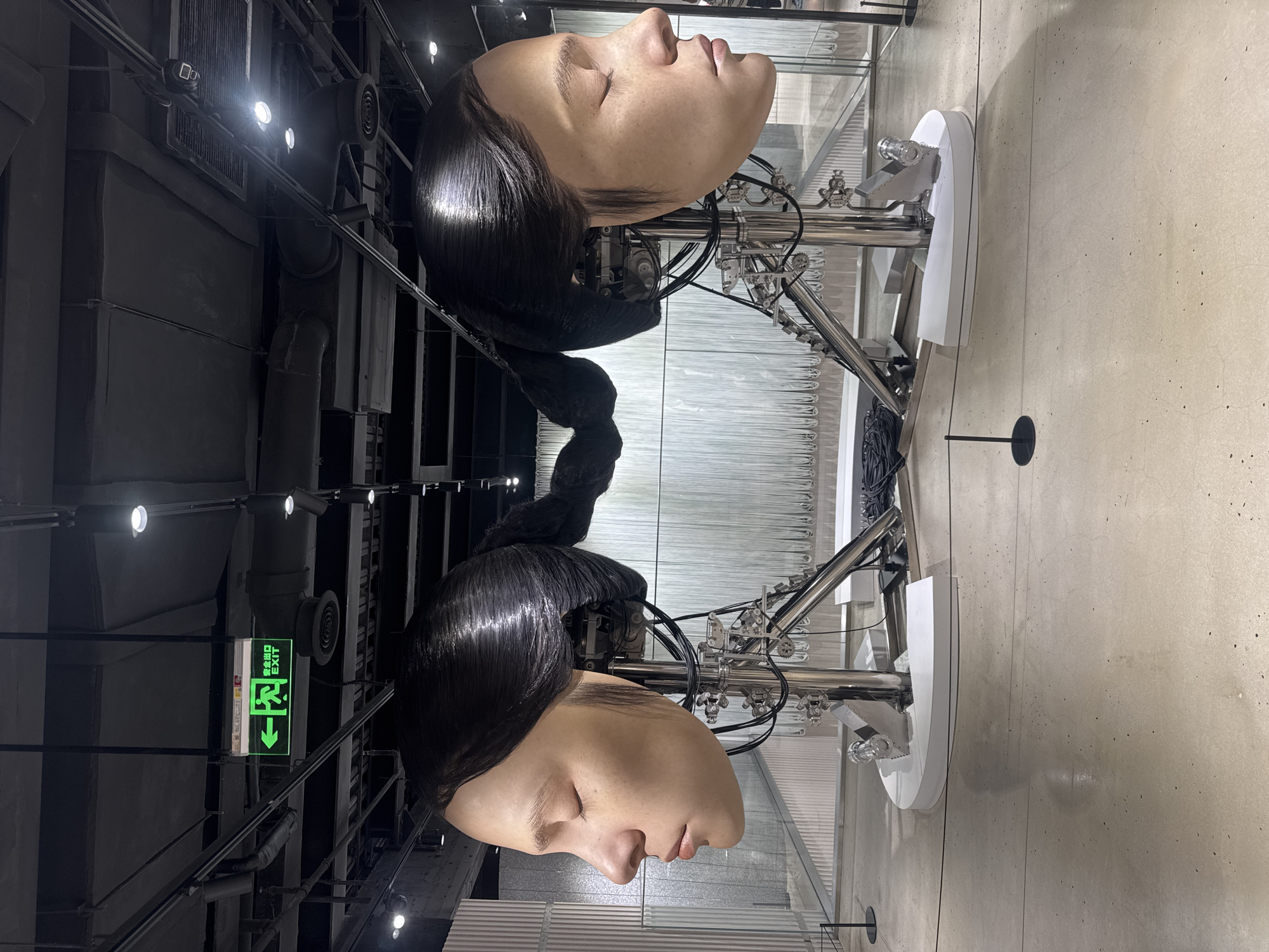 The image shows a modern art installation in a well-lit exhibition space. Two large, realistic robot heads, sculpted to resemble the profile of a young woman with her eyes closed and dark hair pulled back into a braided ponytail, are positioned facing away from each other. They are mounted on complex metallic robotic armatures that are attached to white, curved pedestals. Thin cables and wires are visible, connecting the heads to the armatures. In the background, a wall of vertical, translucent panels creates a waterfall-like effect. Above, the ceiling is dark, with exposed pipes, vents, and track lighting. A green exit sign with an illuminated figure and Chinese characters is visible hanging from the ceiling. The floor is a polished concrete surface. This art installation appears to be located in the Power Station of Art in Shanghai, China, a prominent contemporary art museum.