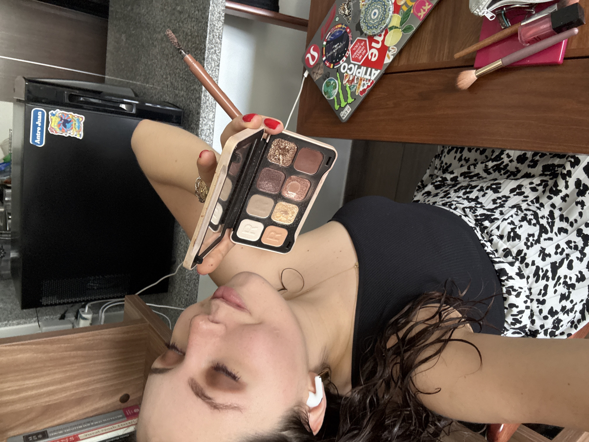 The image captures a person, likely a woman, lying down indoors, viewed from above. She has dark, wavy hair and is wearing a black ribbed top and a black and white patterned skirt. Her eyes are closed, and she holds an open eyeshadow palette featuring various shimmer and matte shades in her right hand, along with a brown-handled makeup brush. Red nail polish is visible on her fingers, and she wears a ring on her left hand. White wireless earbuds are in her ears, and a delicate necklace is visible around her neck.
The setting appears to be a casual personal space. To the left, a black mini-fridge with a blue sticker reading "Antro Juan" is visible. Below her head, books are stacked on a wooden surface, with "Yuval Noah Harari" clearly legible on one spine. To her right, an open silver laptop, heavily adorned with colorful stickers, sits on a wooden table. Some legible text on the stickers includes "ATIPICO", "NO ME HABLES, ME DISTRAIGO FACIL", and "ESN". Next to the laptop, several other makeup items, including a pink compact, a makeup brush, and a lip gloss, are arranged. The overall scene suggests a moment of relaxation or self-care in a lived-in environment in Mexico City, Mexico.